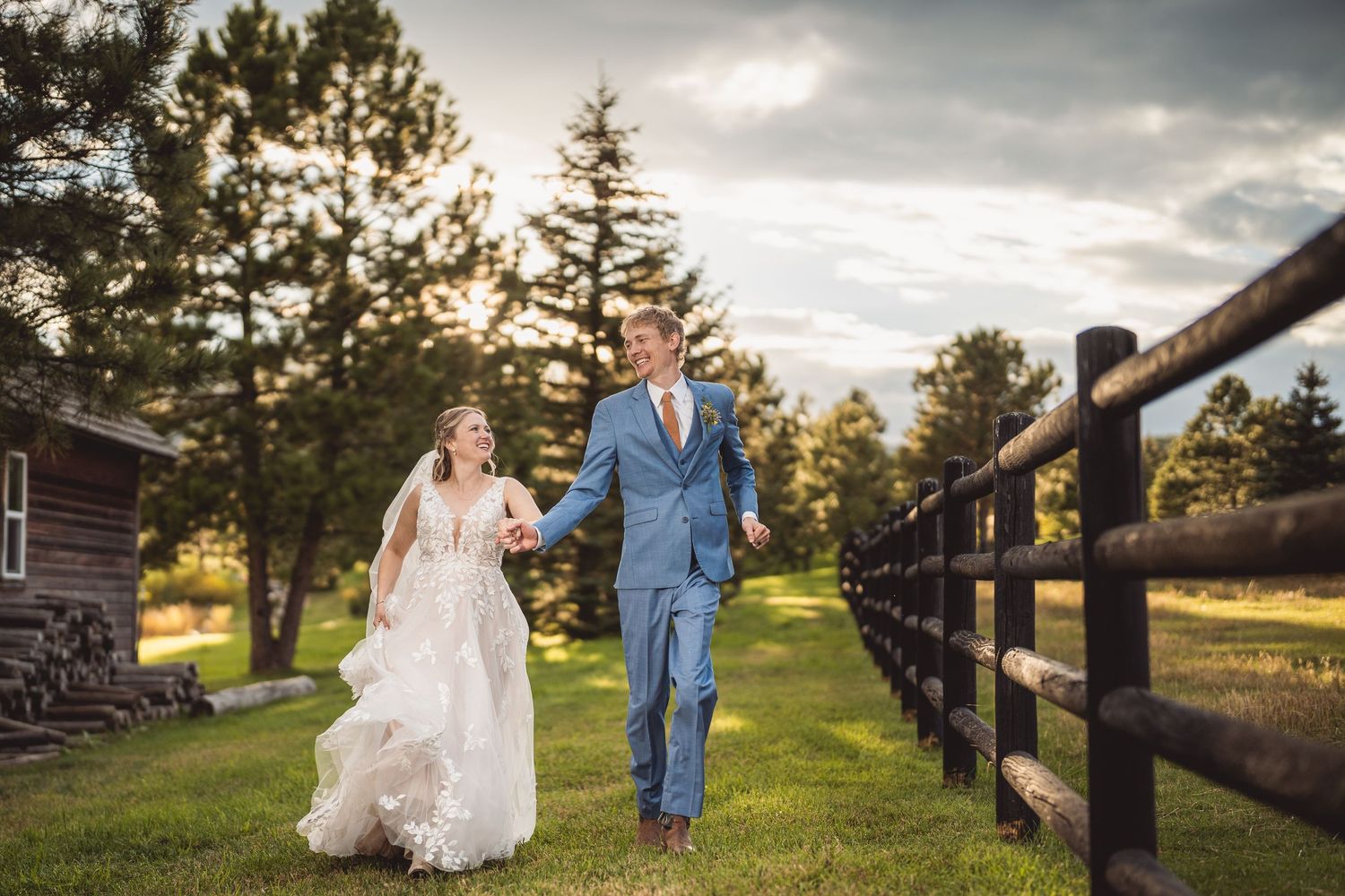 Bride and groom walk hand-in-hand along a wooden fence at golden hour, surrounded by lush green trees.