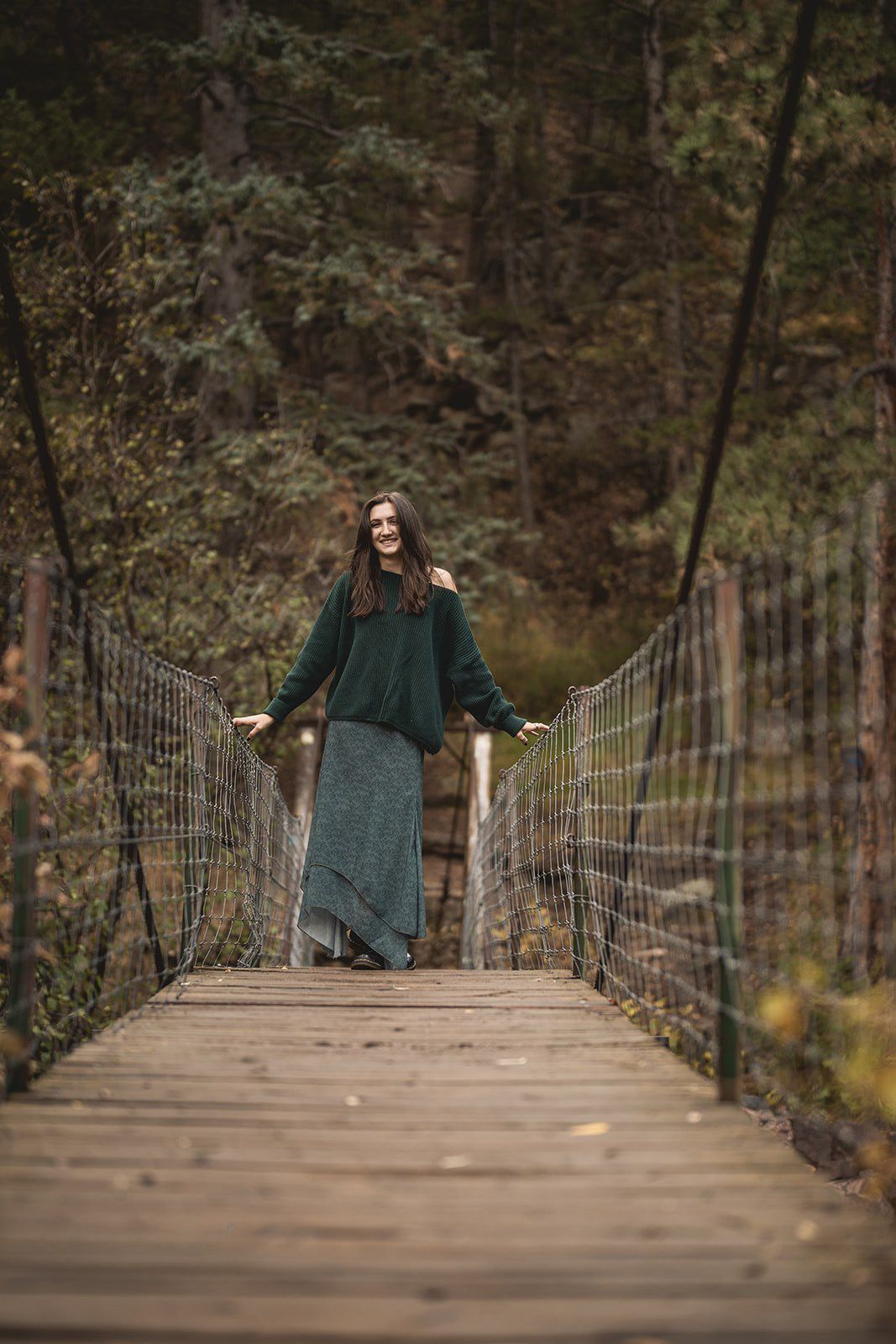 Smiling woman in a green sweater and gray skirt standing on a wooden suspension bridge in autumn forest.
