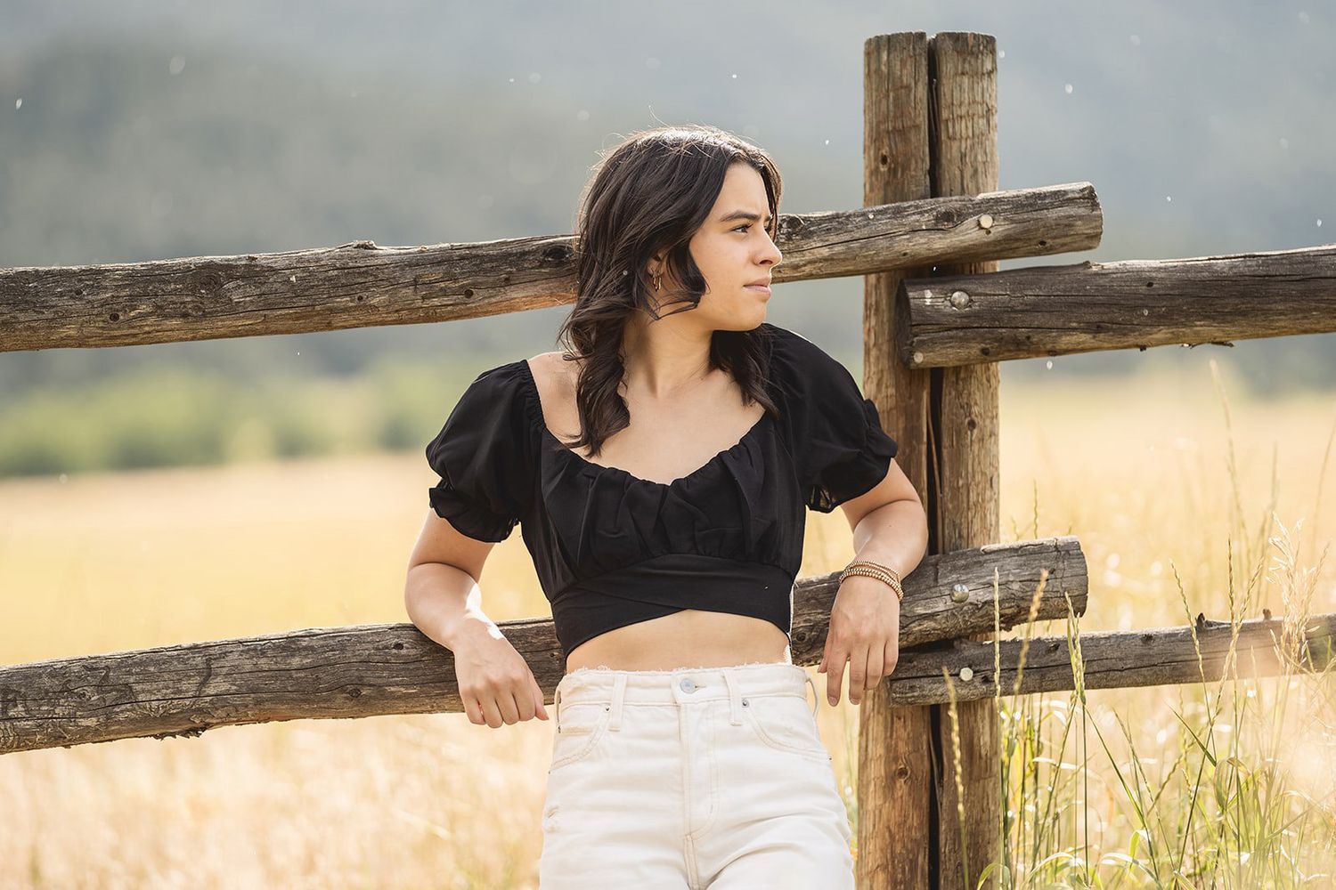 Young woman in black crop top and white jeans posing by a rustic wooden fence in a sunny field.