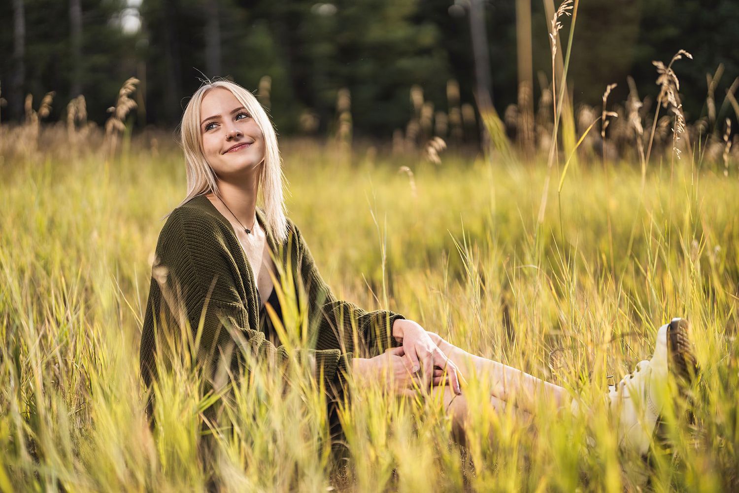 Blonde woman sitting in tall golden grass, smiling upward in a sunlit field.