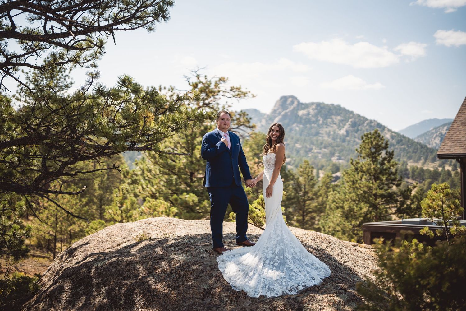 Bride and groom stand on a rocky overlook surrounded by pine trees and mountain views in Colorado.