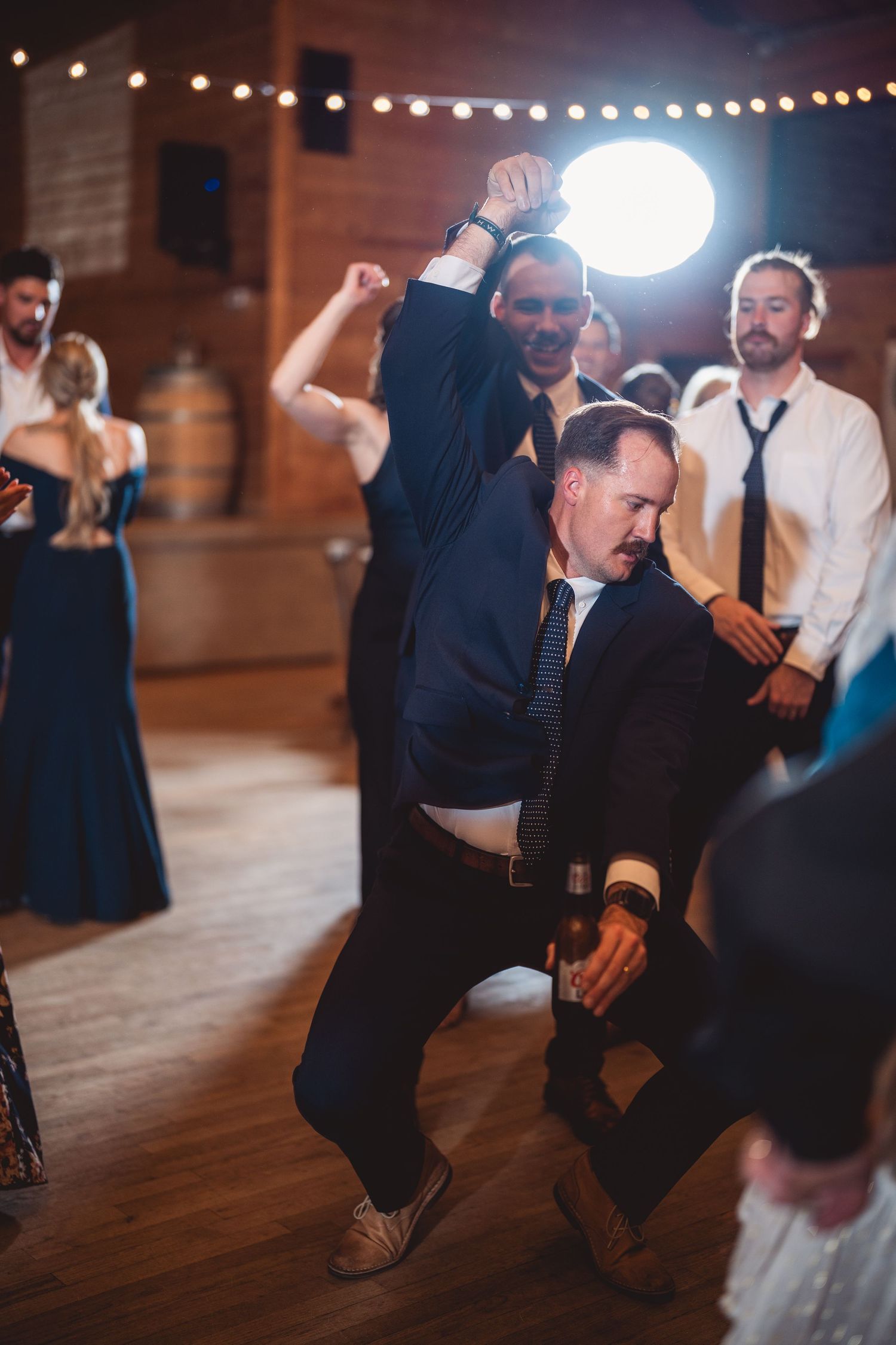 Groomsmen celebrate and dance energetically at a rustic barn wedding reception with string lights.