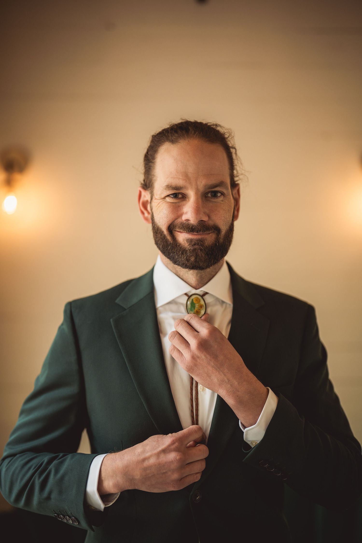 Bearded groom in dark green suit adjusts bolo tie while smiling warmly in softly lit room.