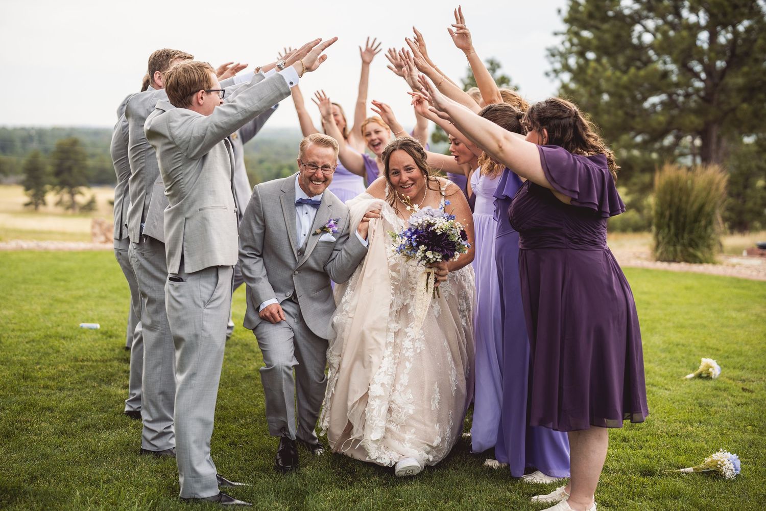 Wedding party celebrating with arms raised outdoors in purple and grey attire.