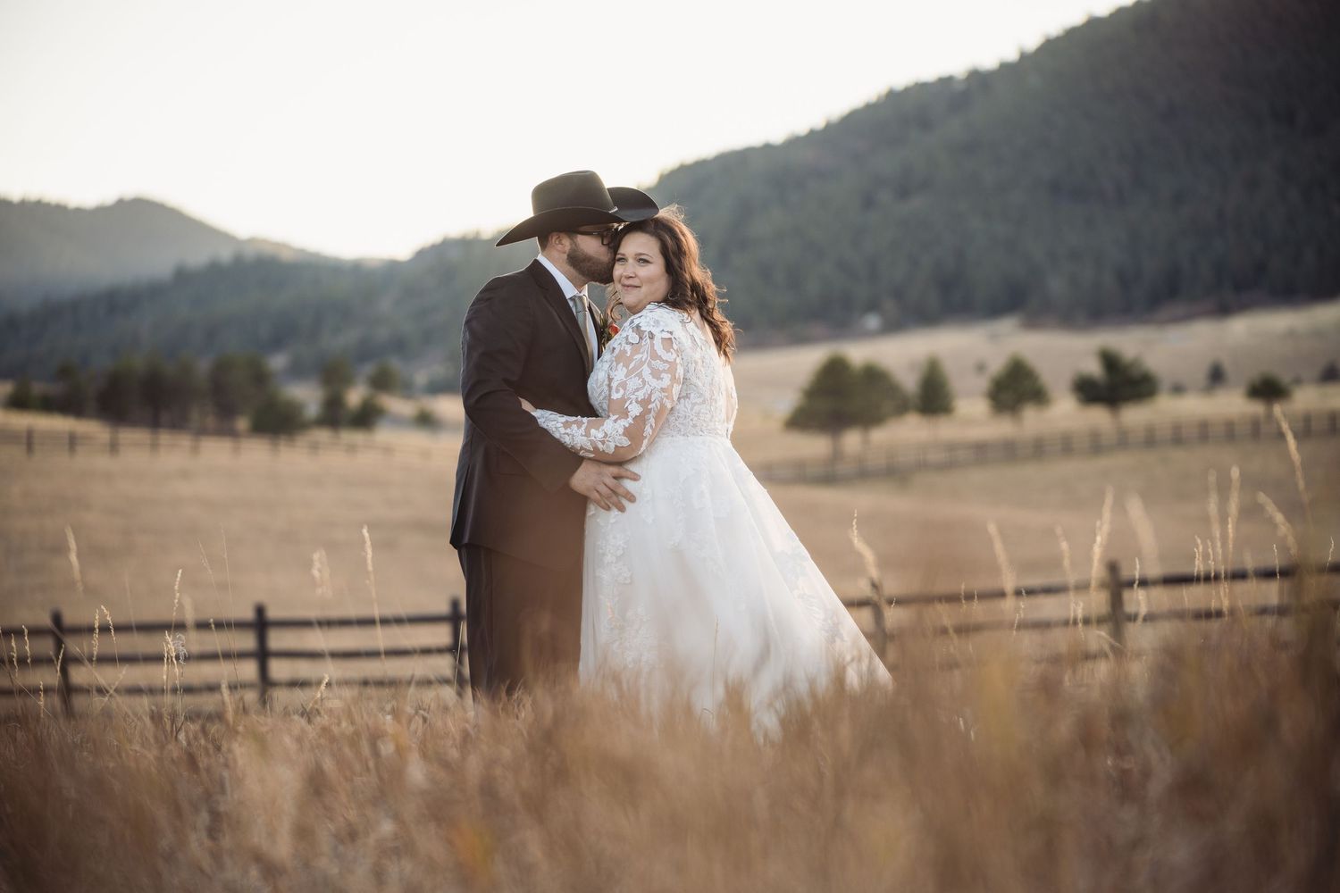 A romantic embrace in a rustic field with mountain views and wooden fencing at golden hour.