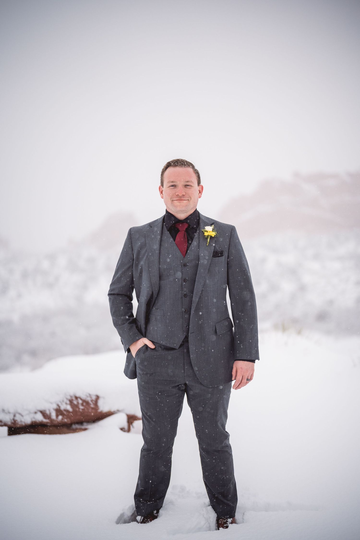 Groom in charcoal suit poses in snowy winter landscape.