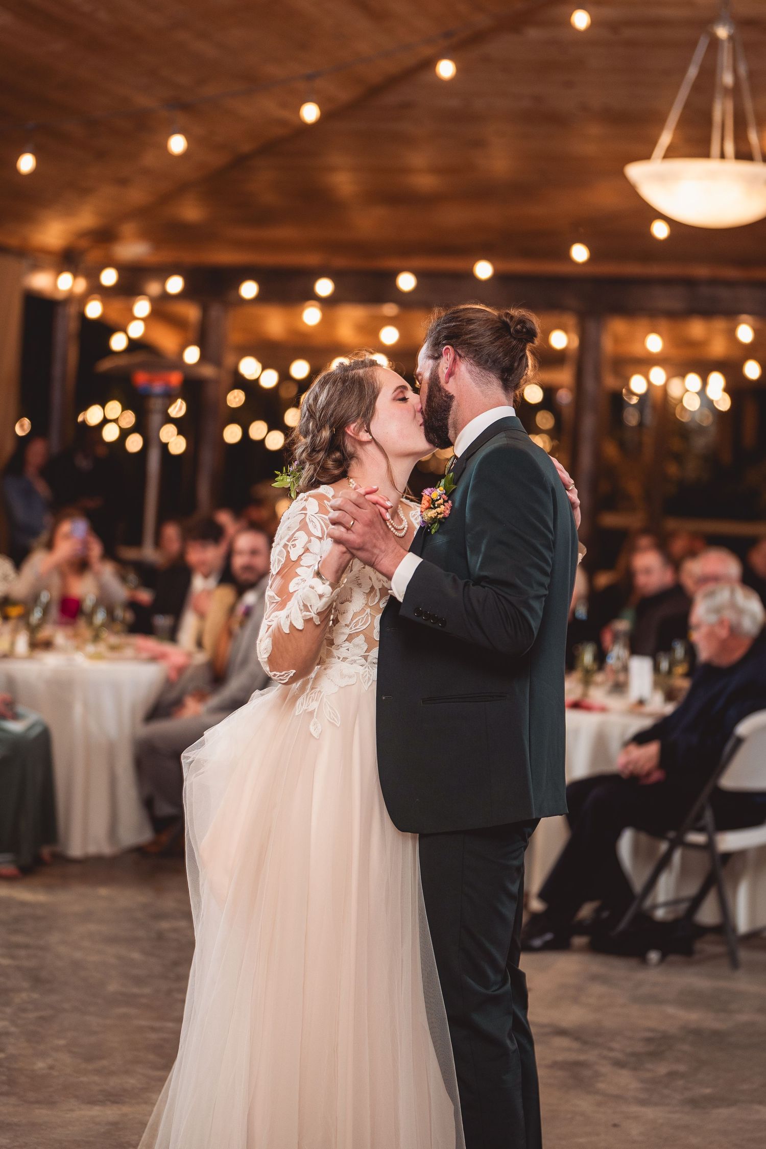 Newlyweds share their first dance in a rustic reception hall decorated with warm string lights overhead.