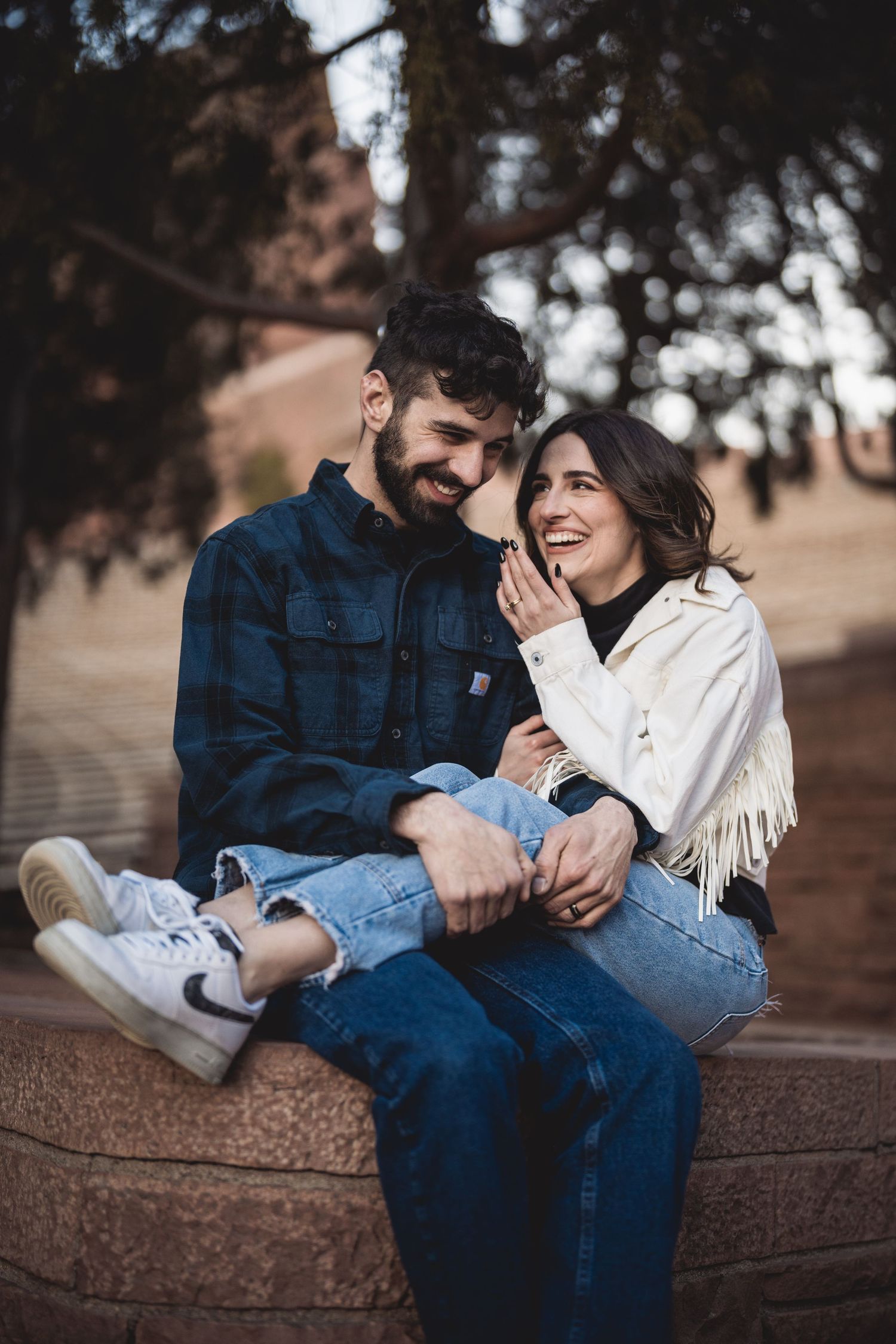Happy couple sitting outdoors, woman in white fringe jacket laughing with bearded man in blue flannel shirt and jeans.