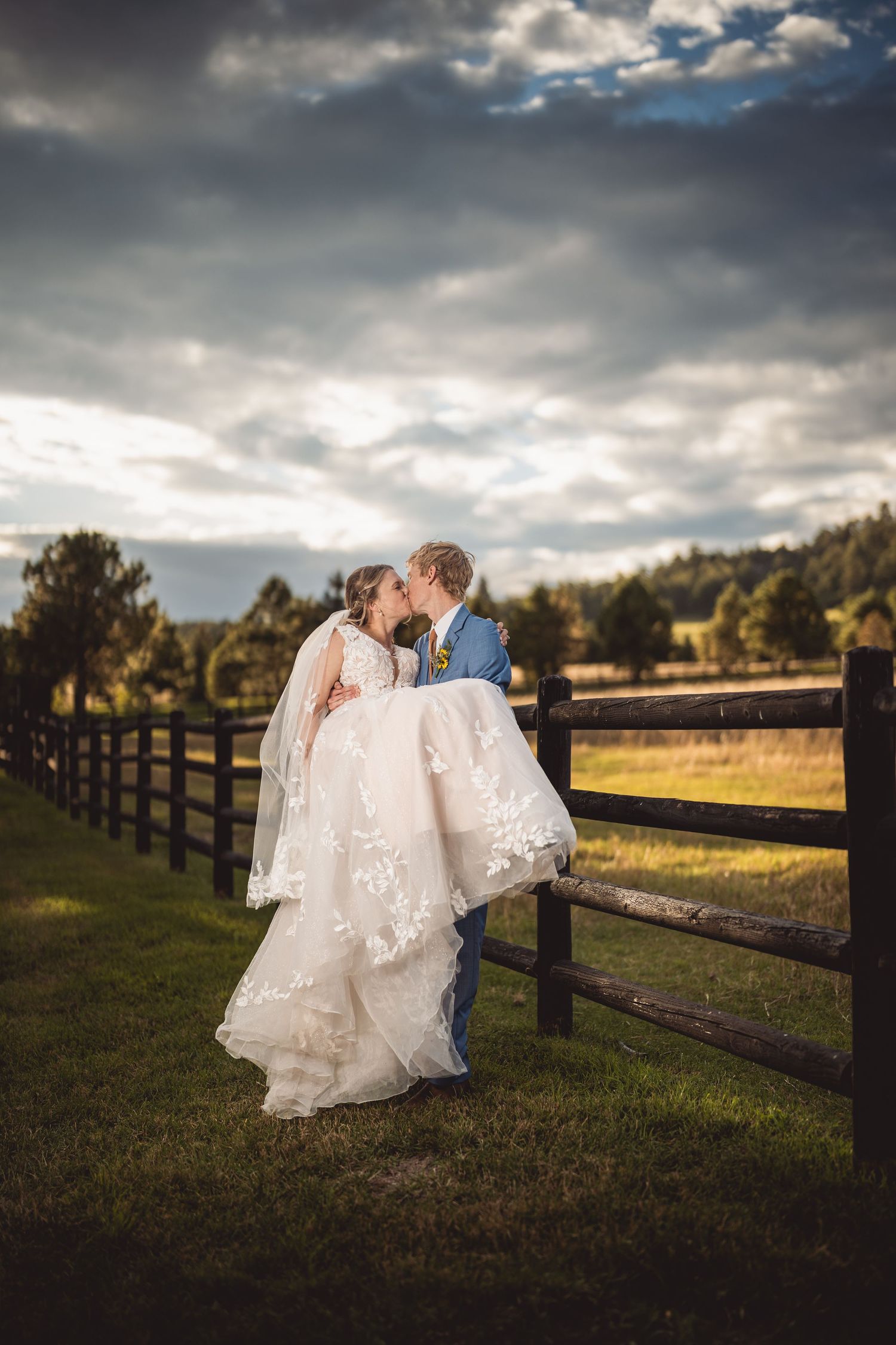 A bride and groom share a romantic kiss by a rustic wooden fence at a countryside wedding venue under dramatic cloudy skies.
