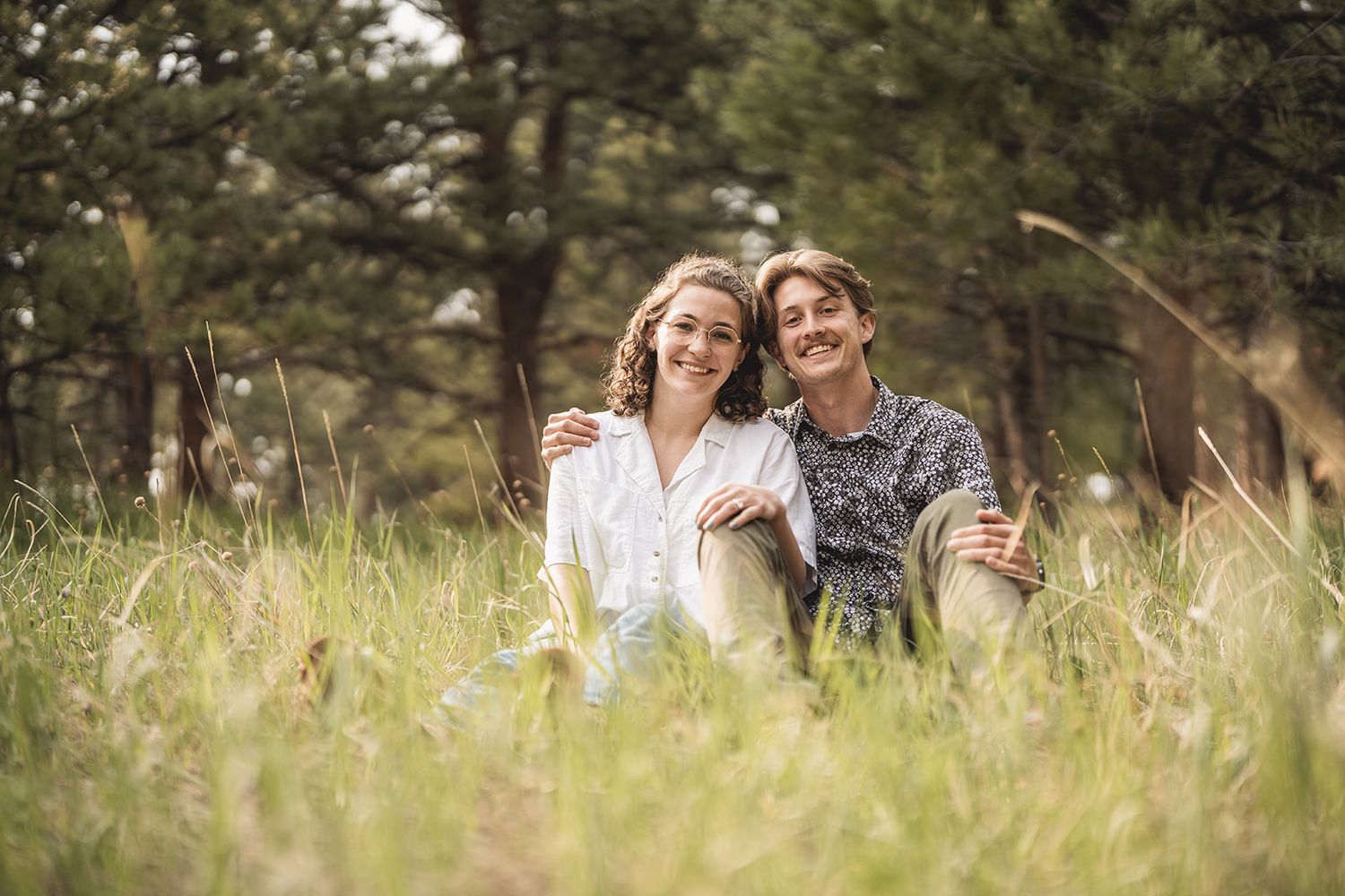A smiling couple sits together in a lush green meadow surrounded by trees, enjoying a relaxed outdoor moment.