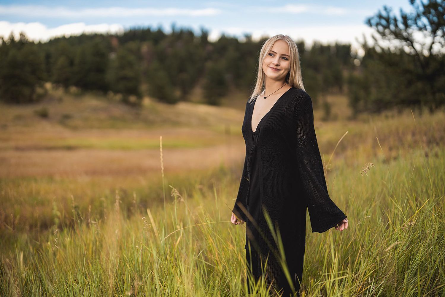Blonde young woman in black maxi dress standing in tall grass field with forest background.