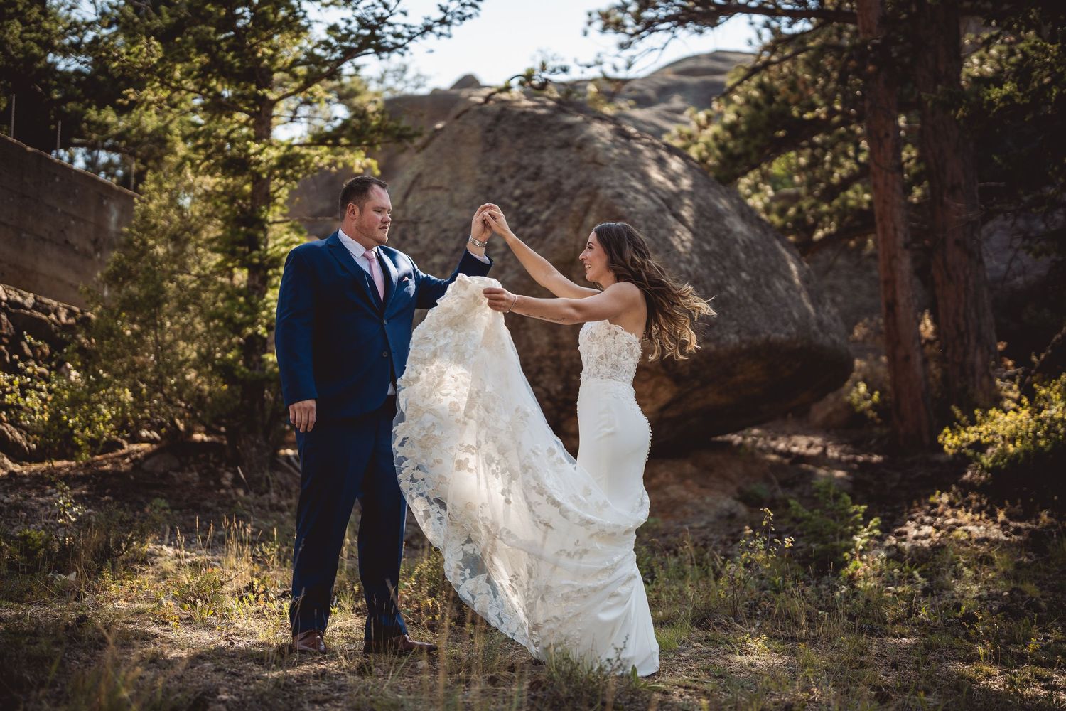 A couple dances and twirls in a white dress and navy suit among pine trees.