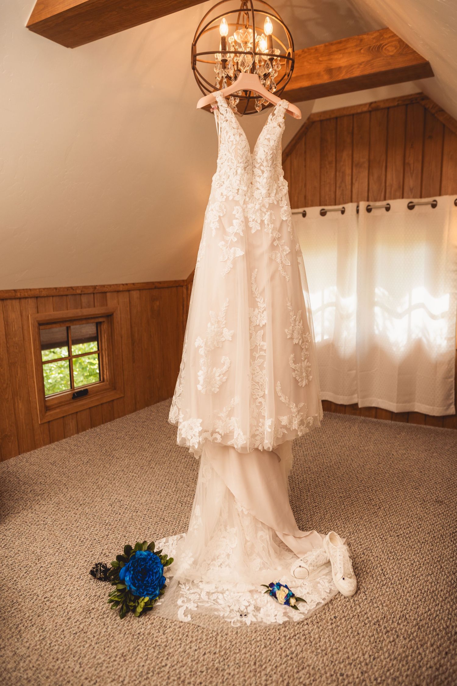 White lace wedding dress hanging from wooden ceiling with chandelier in rustic room.