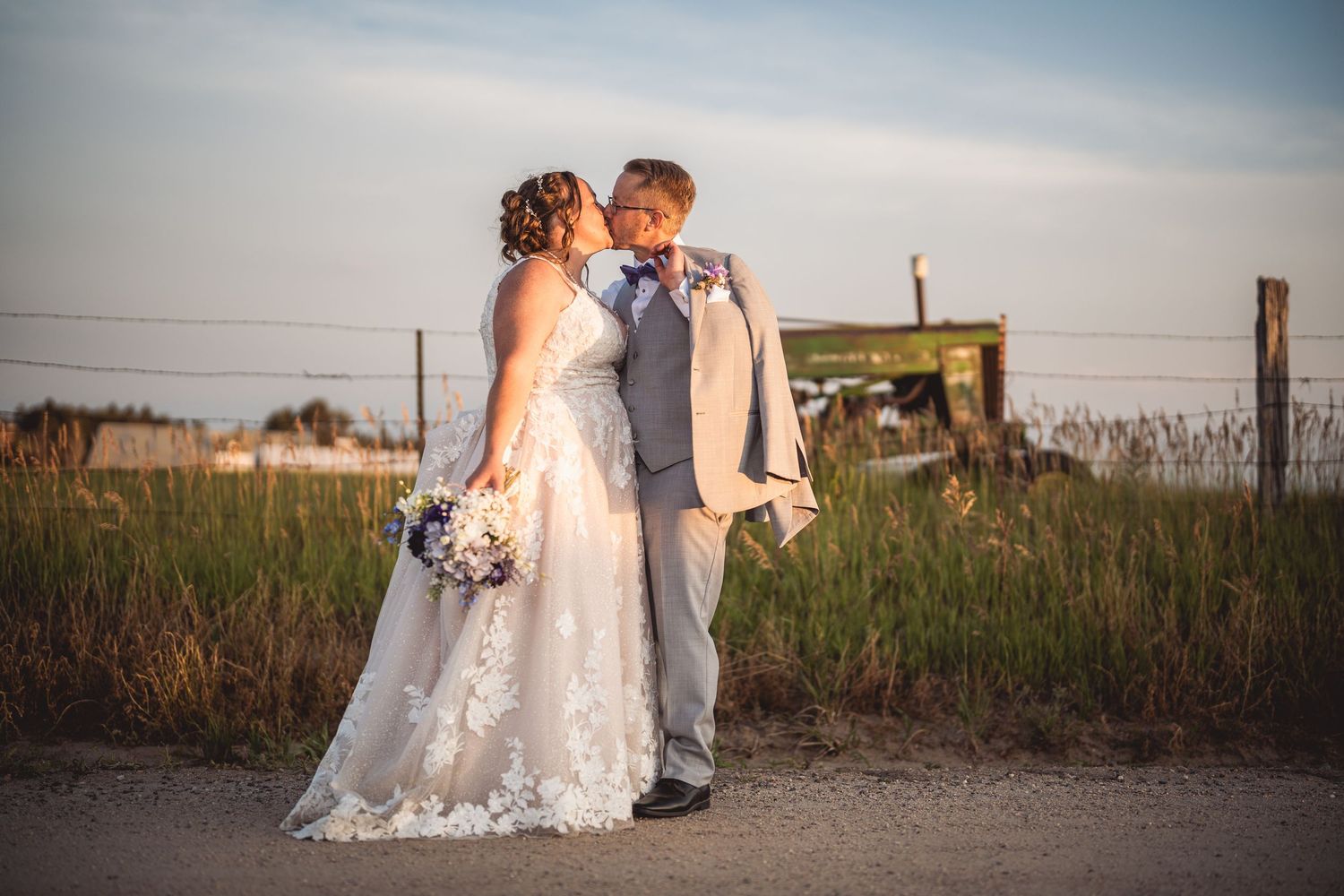 Couple poses together on rural path with lace wedding dress flowing in evening light.