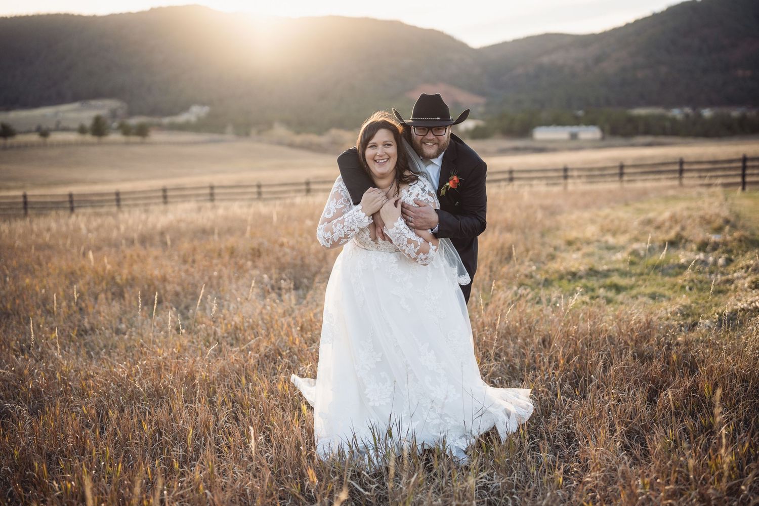 Newlyweds embracing in rustic field at sunset with mountains in background.