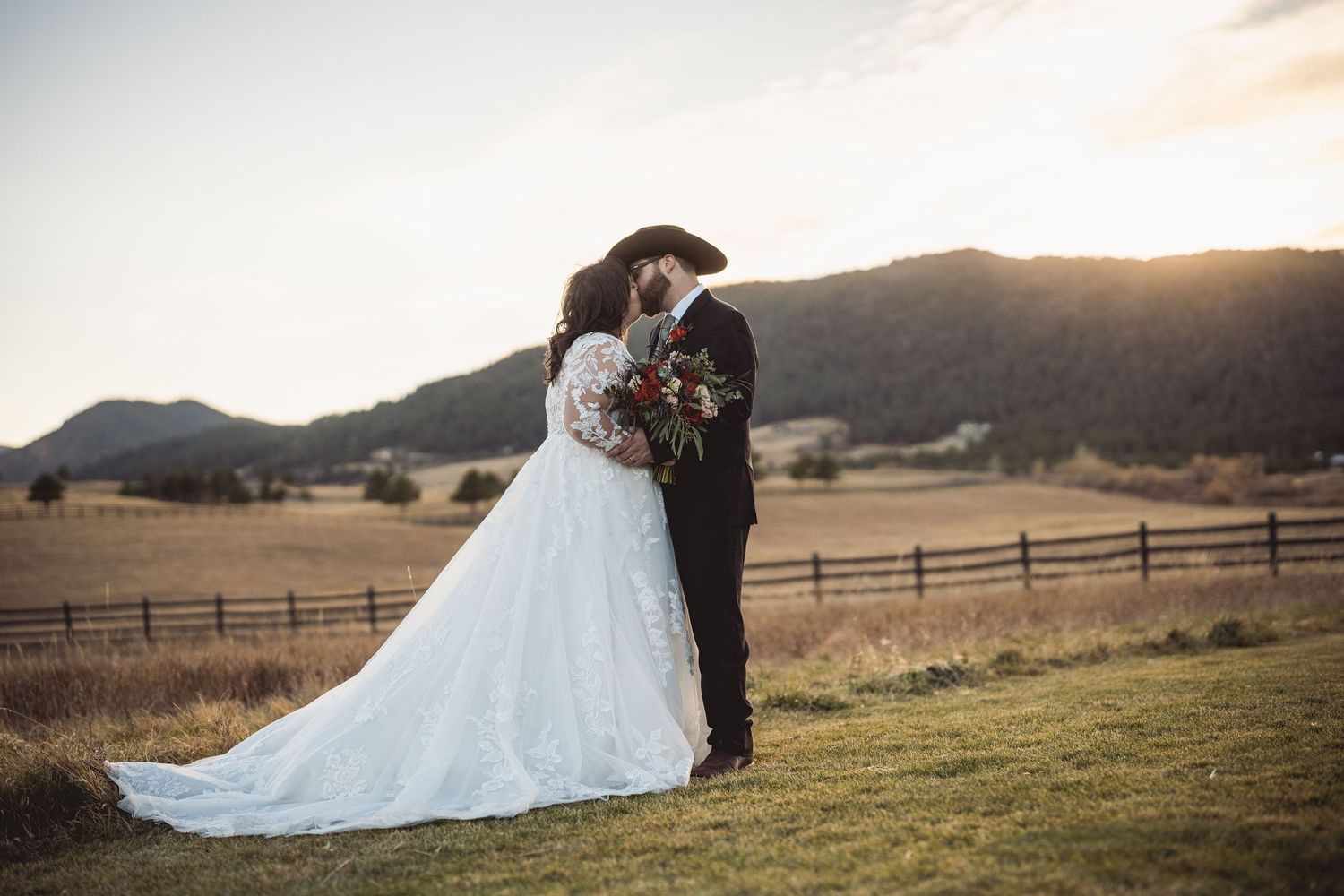 Romantic sunset wedding portrait against rustic wooden fence and mountain backdrop showing flowing white dress.