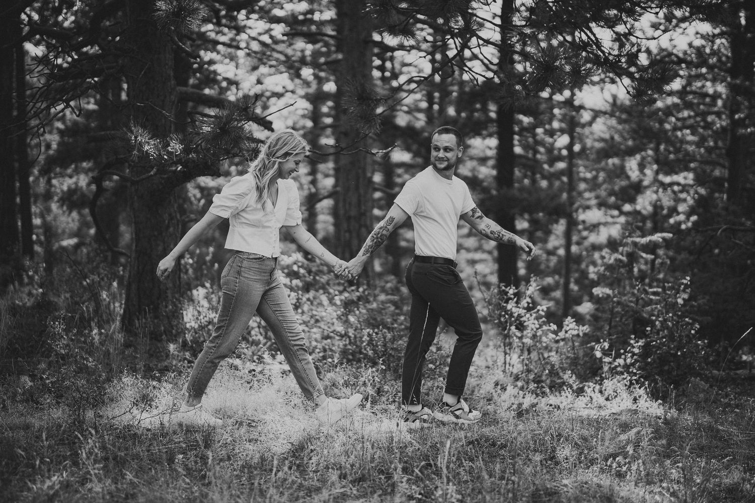 Black and white photo of a couple holding hands and walking through a forest clearing surrounded by tall trees.