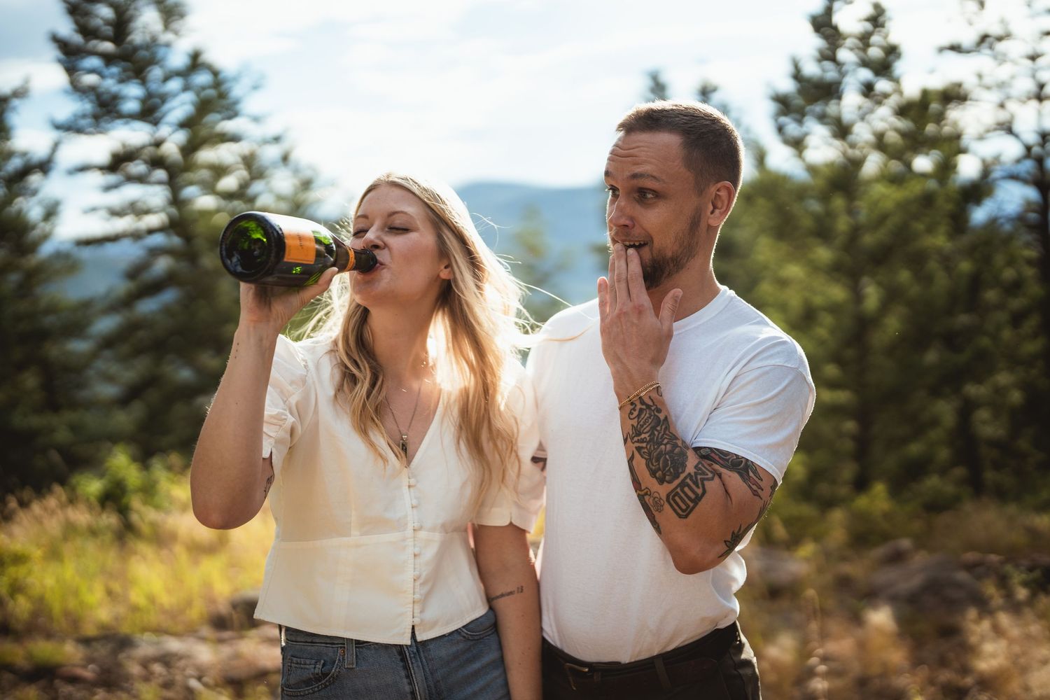 Couple outdoors among pine trees, woman drinking from wine bottle, tattooed man beside her in white shirt.