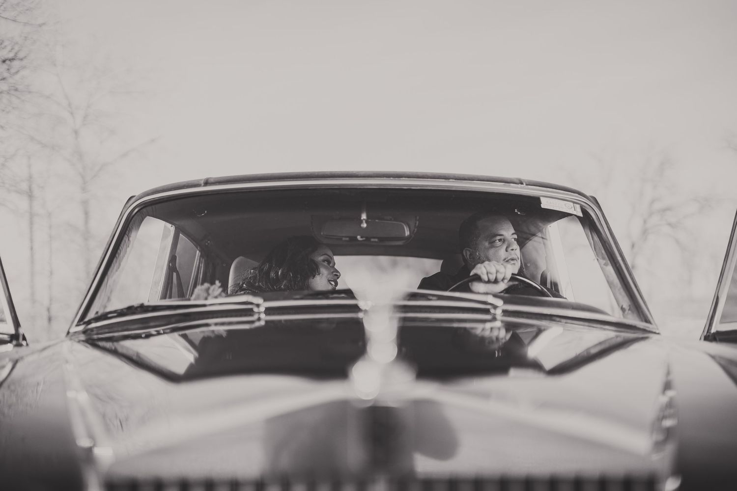 Black and white photo of two people sitting in a vintage classic car, viewed from the front windshield.