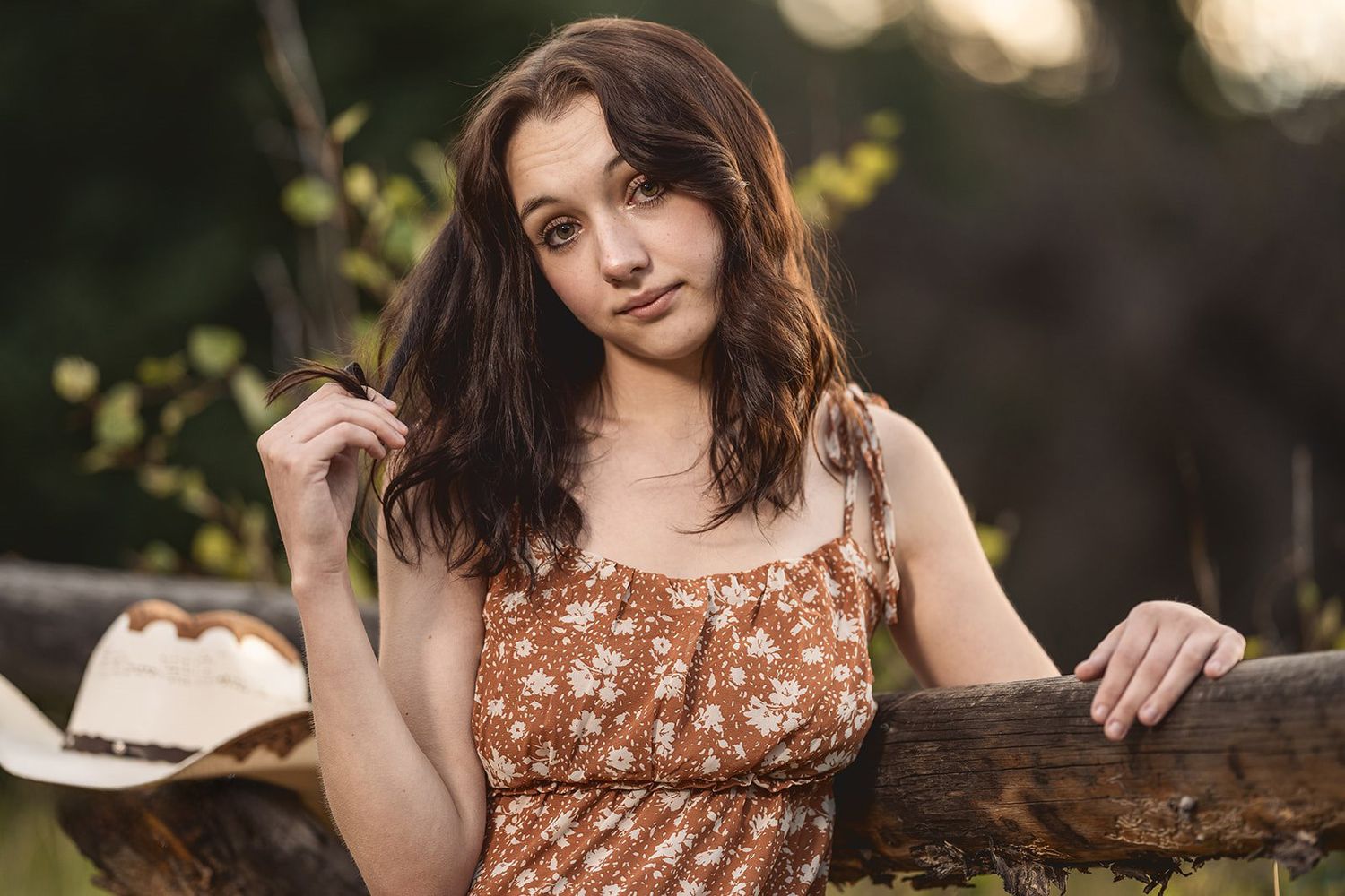 Young woman in floral sundress leaning on a rustic wooden fence in golden hour outdoor portrait photography.