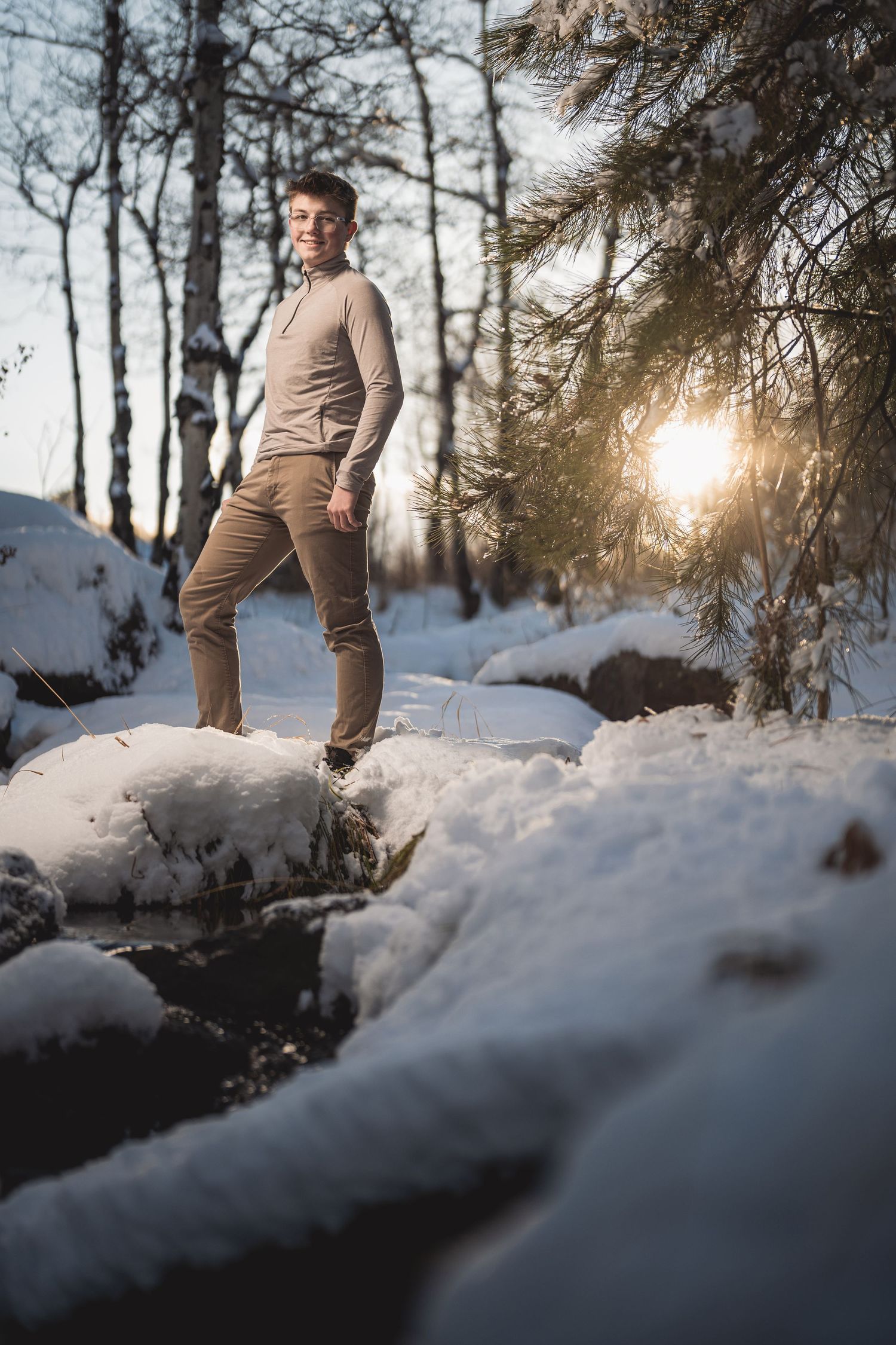 Teen boy standing on snowy rocks in a winter forest with golden sunset light through pine trees.