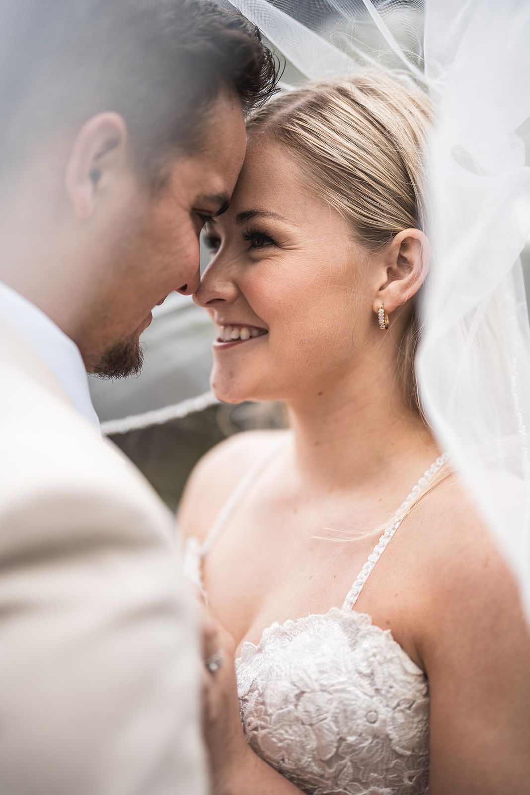 Smiling bride in lace dress and groom in tan suit share an intimate moment under a wedding veil.