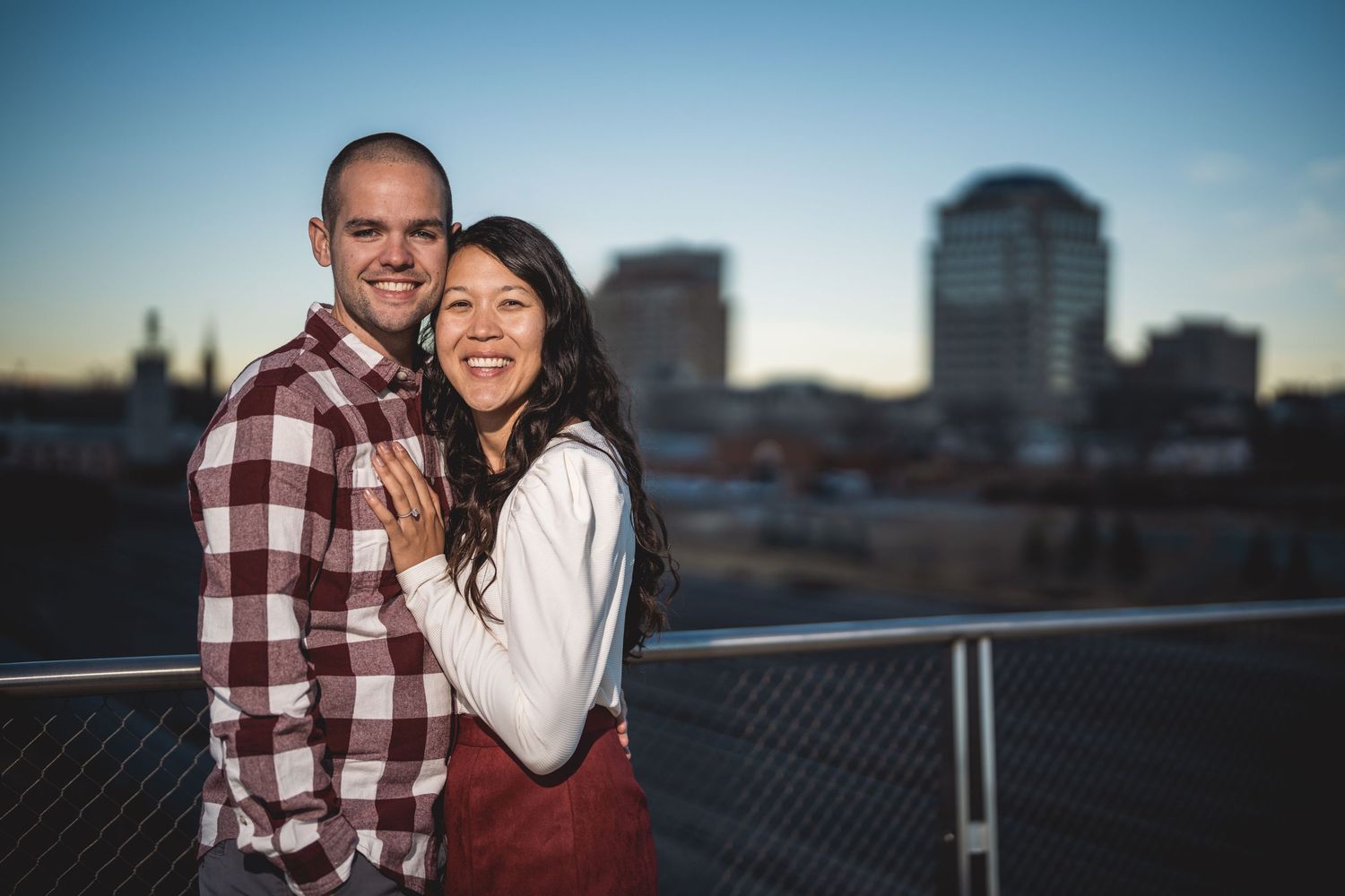 Smiling couple hugging on a rooftop with city skyline at dusk in the background.