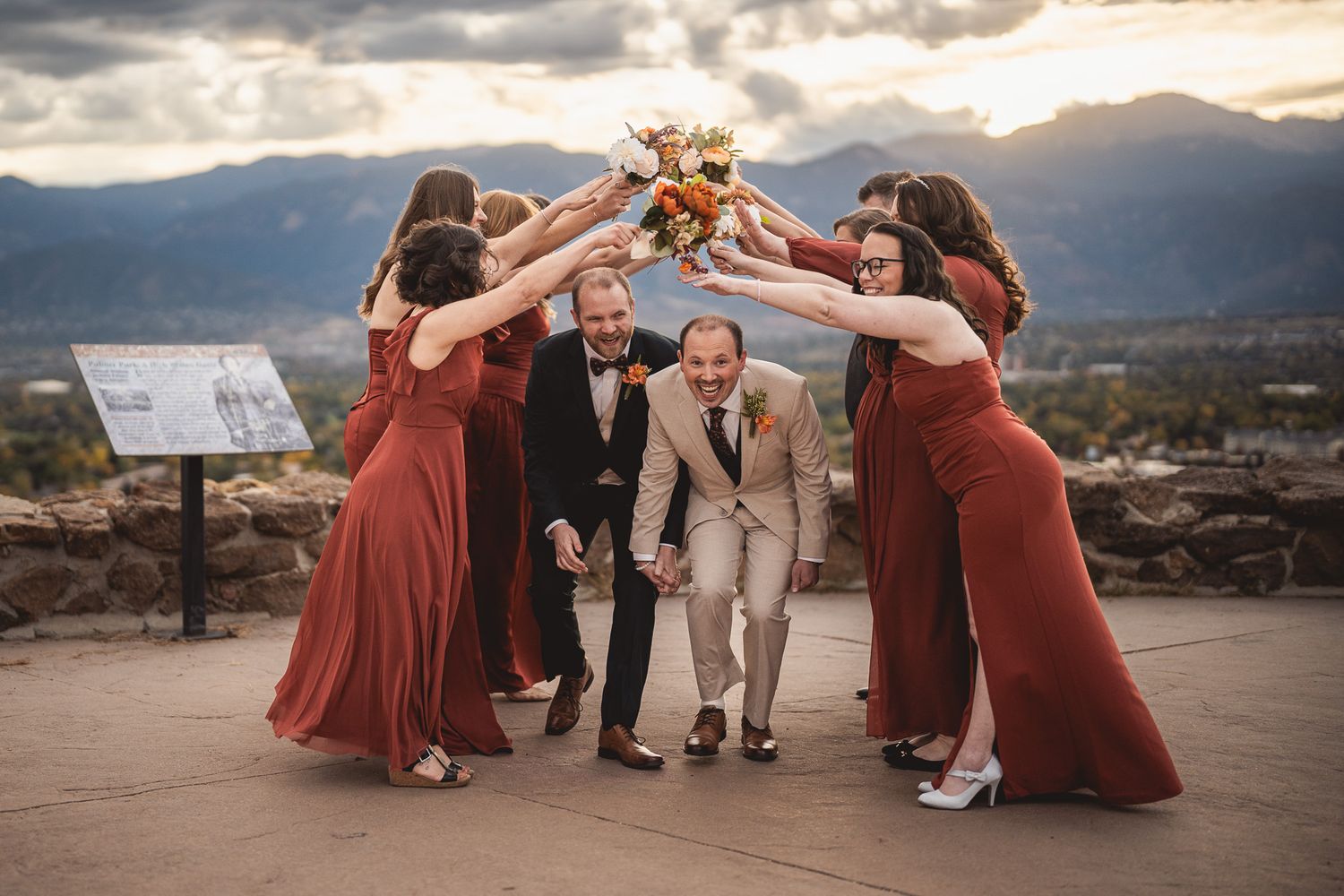 Wedding party celebrates under an arch of raised hands at a scenic mountain overlook at sunset.