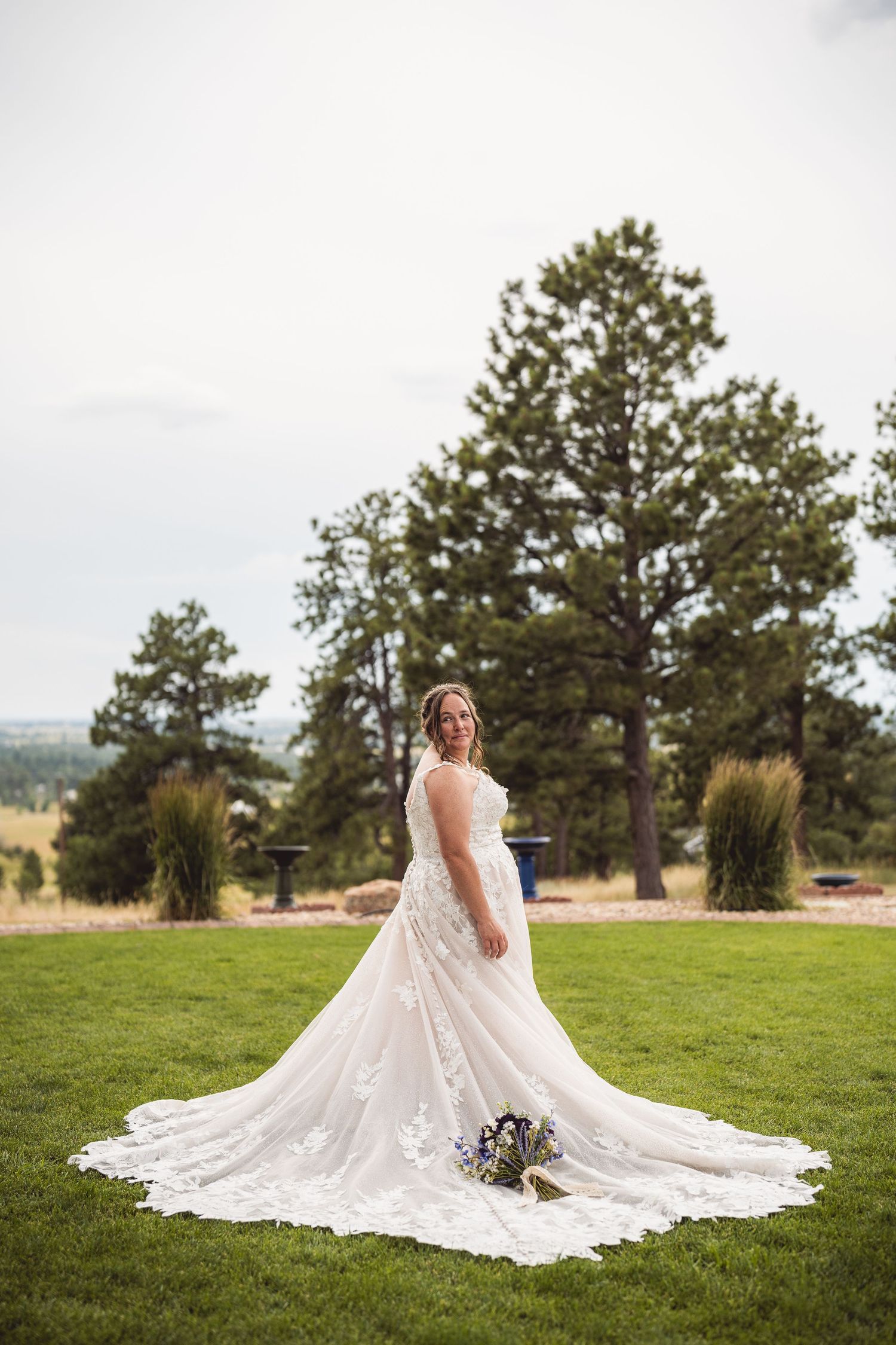 Bride in elegant lace wedding gown with long train holds bouquet on green lawn with pine trees.