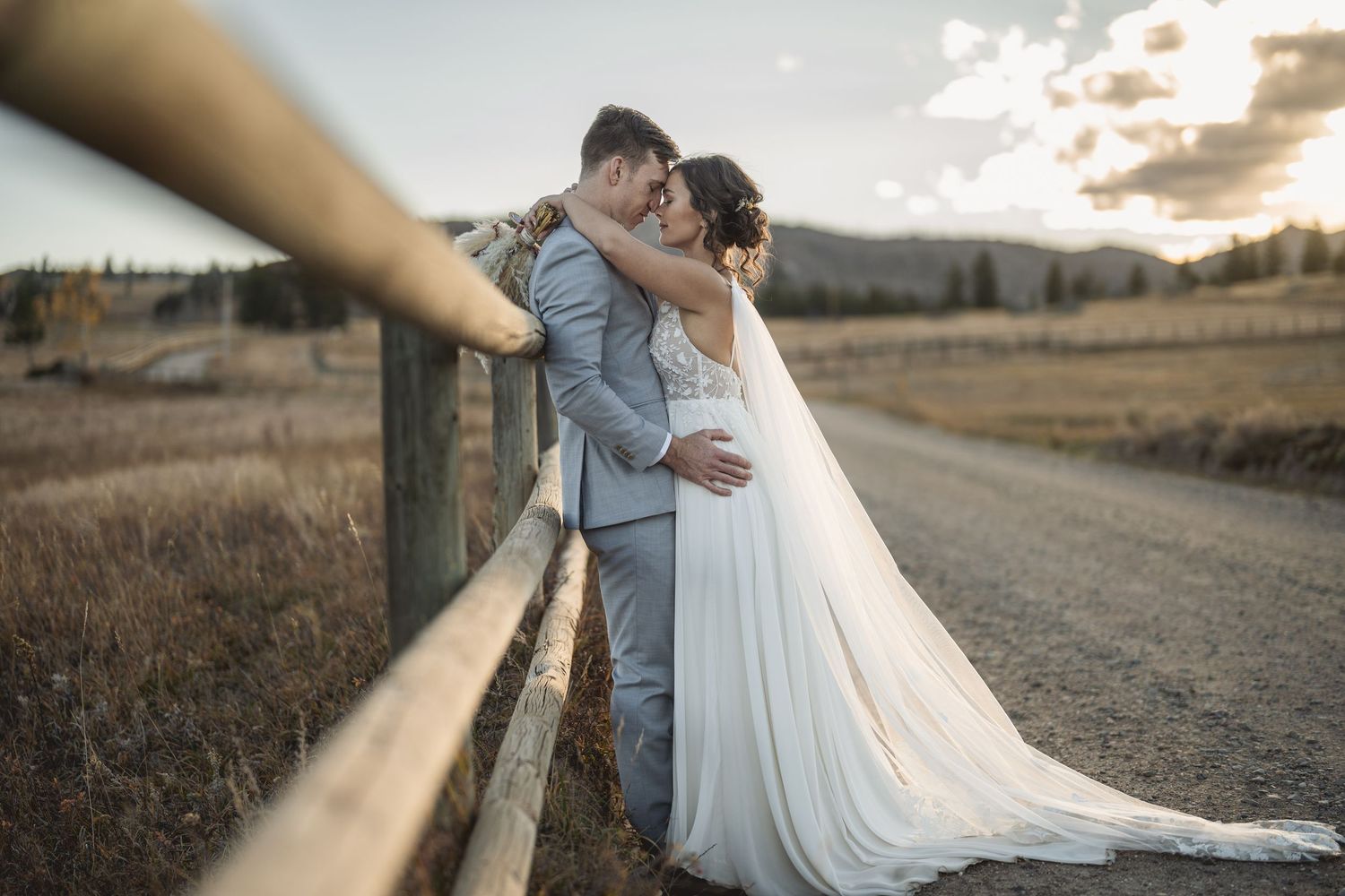 Couple sharing intimate moment on rural path with sunset glow and flowing white dress.