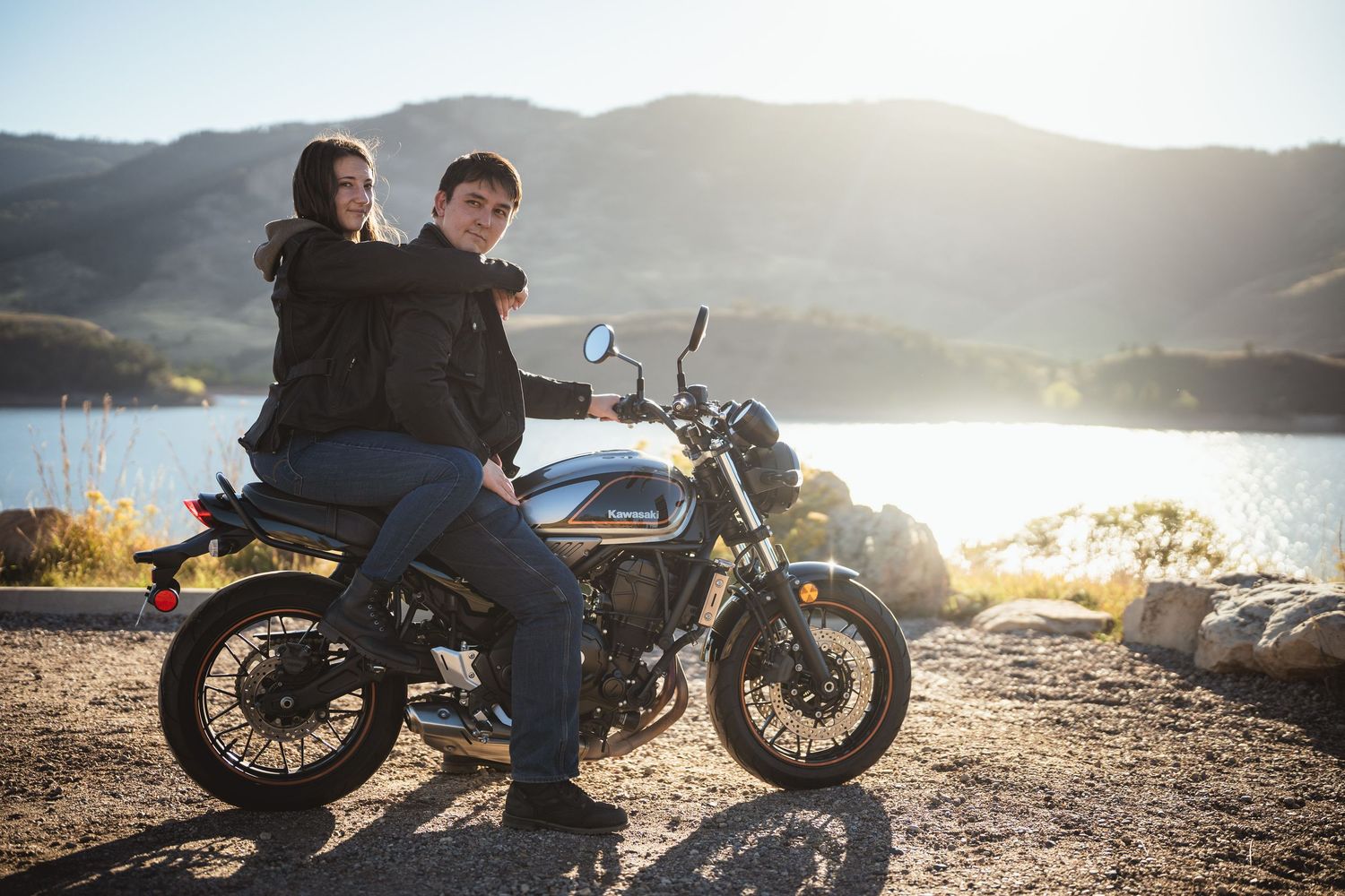 Couple sits together on a vintage motorcycle by a lake at sunset.