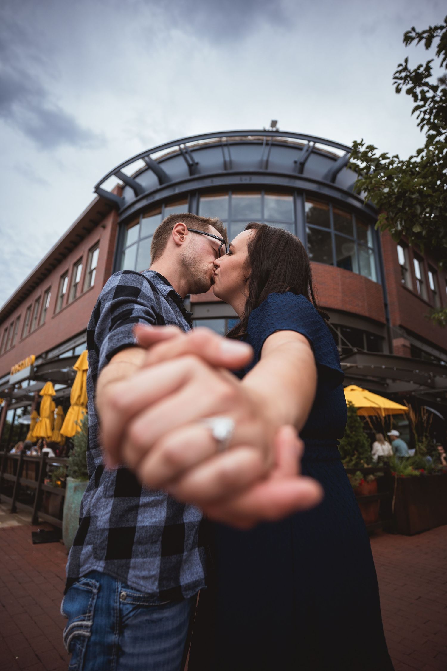 Couple sharing a romantic kiss while holding hands showing engagement ring outside a brick building with yellow umbrellas.