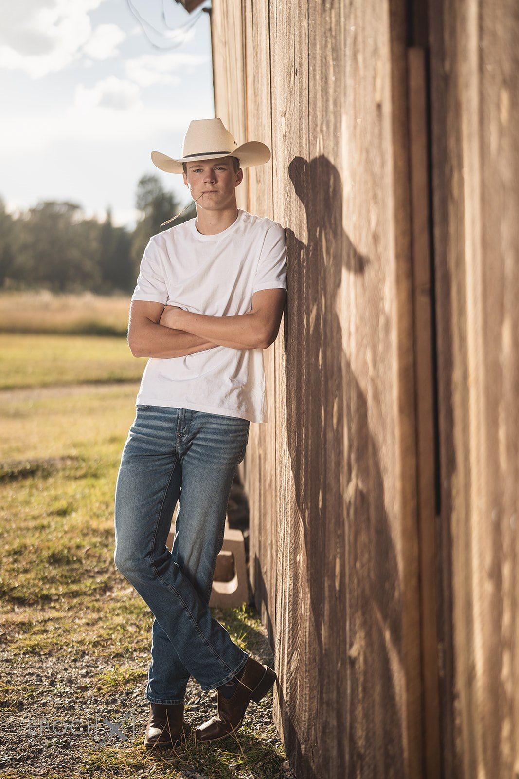 Young man in white t-shirt, jeans, and cowboy hat leaning against rustic wooden barn wall with arms crossed.