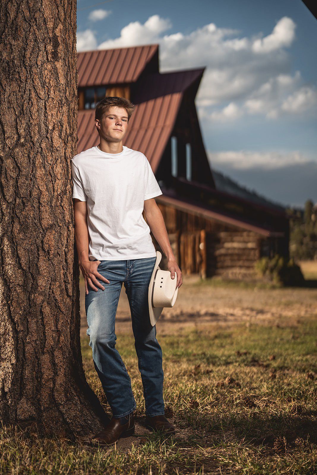 Teen boy in white t-shirt and jeans leaning against large tree holding cowboy hat near log cabin.