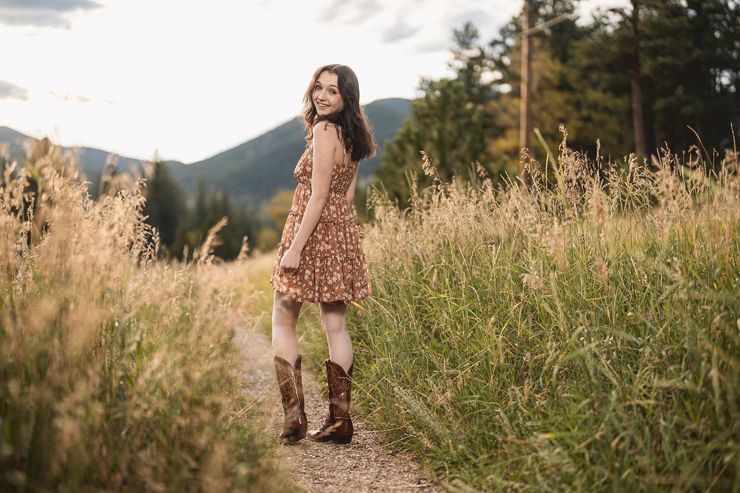 Young woman in floral dress and cowboy boots standing on a mountain trail surrounded by tall grass.
