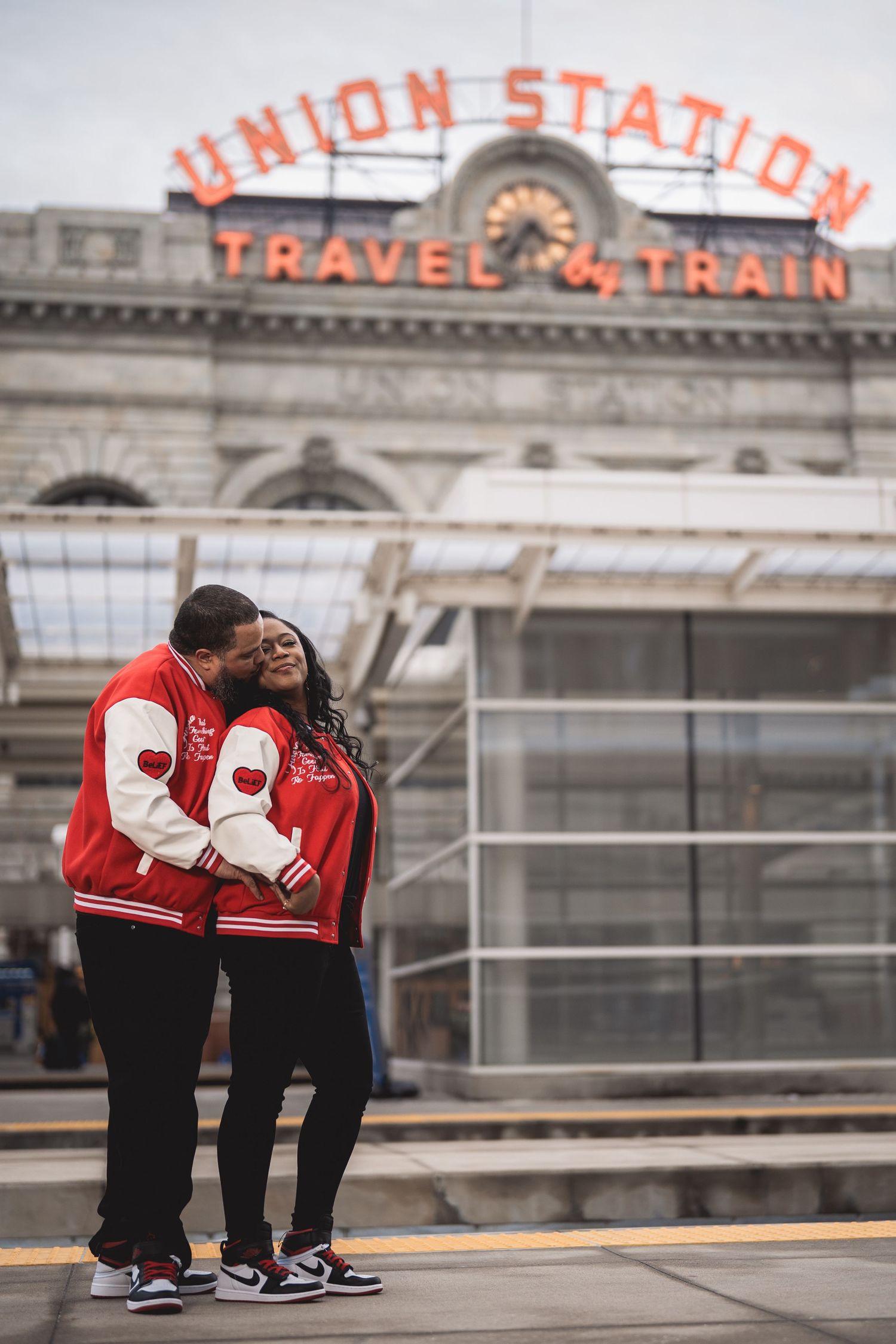 Couple in matching red varsity jackets embracing at Denver Union Station Train Travel sign backdrop.