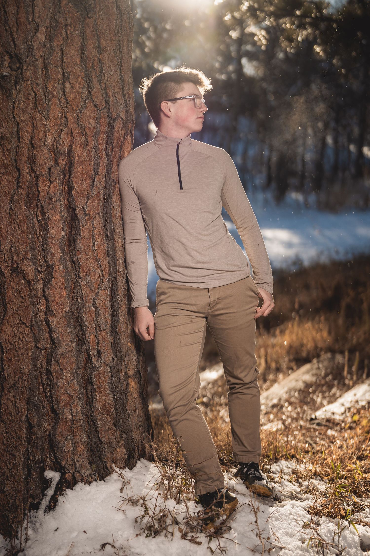 Man in beige outfit leaning against pine tree in snowy winter forest with sunlight filtering through trees.