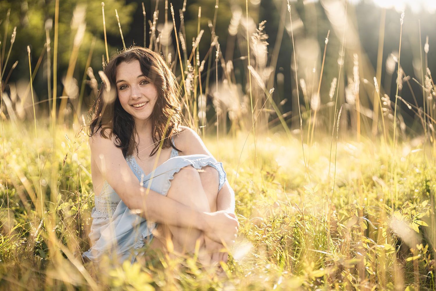 Smiling young woman in light blue dress sitting in sunlit tall grass field during golden hour.