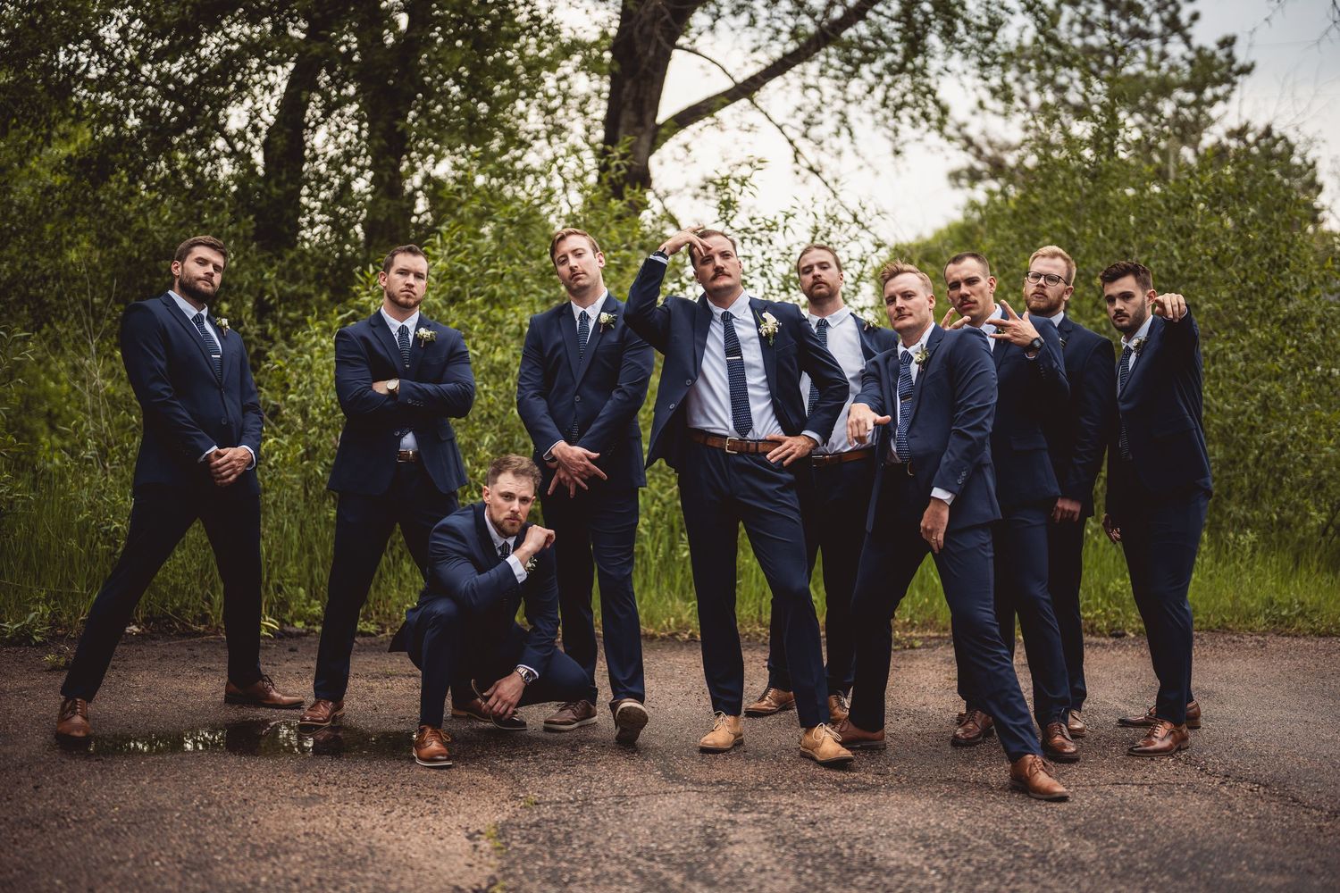 Groomsmen in navy suits pose playfully outdoors on a paved path surrounded by lush green trees.
