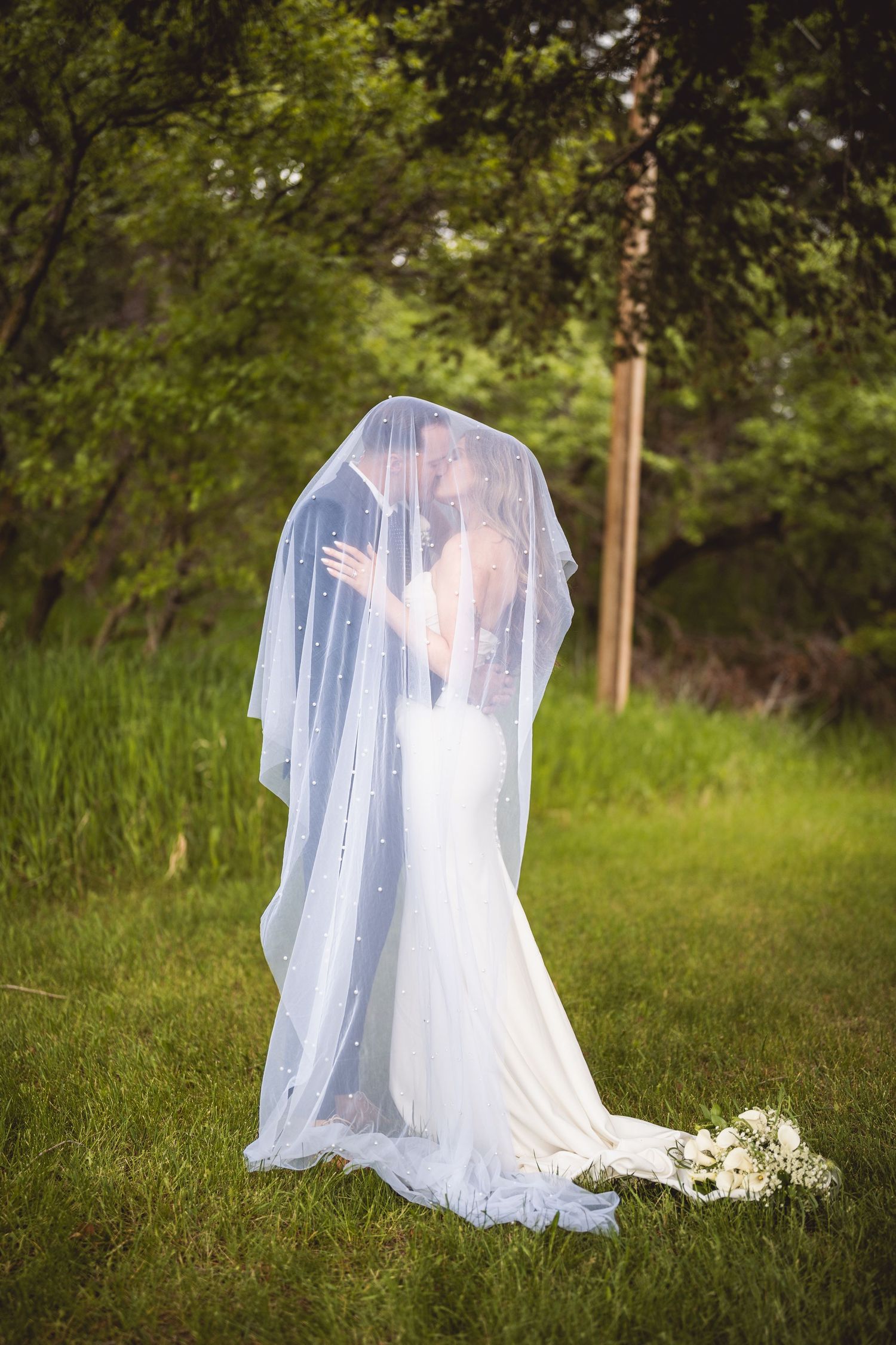 Bride and groom kissing under a flowing white veil in a lush green outdoor setting on their wedding day.