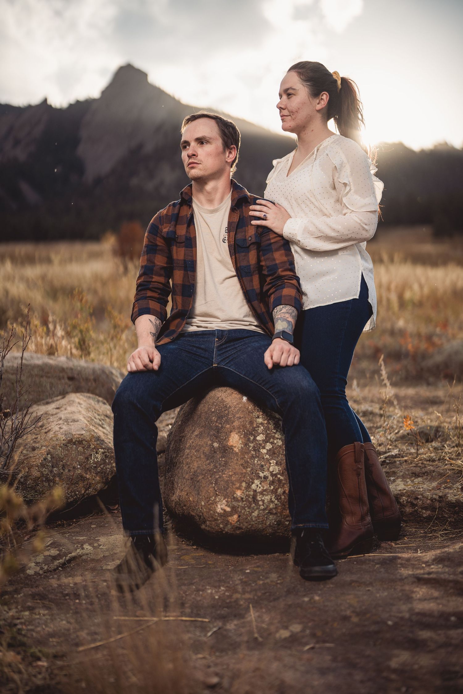 Couple posing outdoors on a boulder with mountain backdrop, man in plaid shirt seated, woman standing behind him.