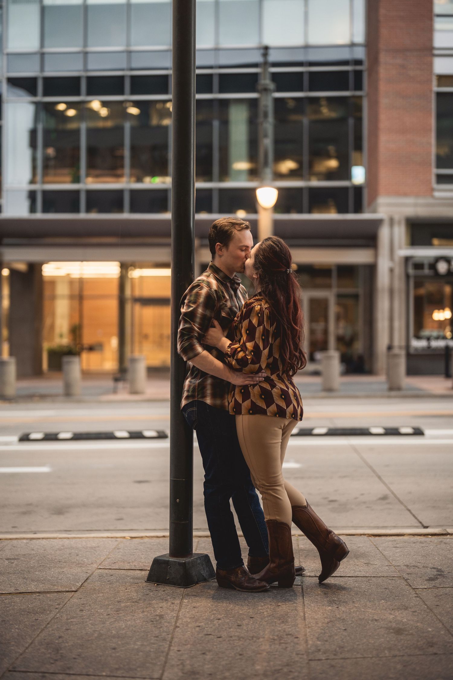 Couple sharing a romantic kiss leaning against a street lamp on an urban sidewalk at dusk.