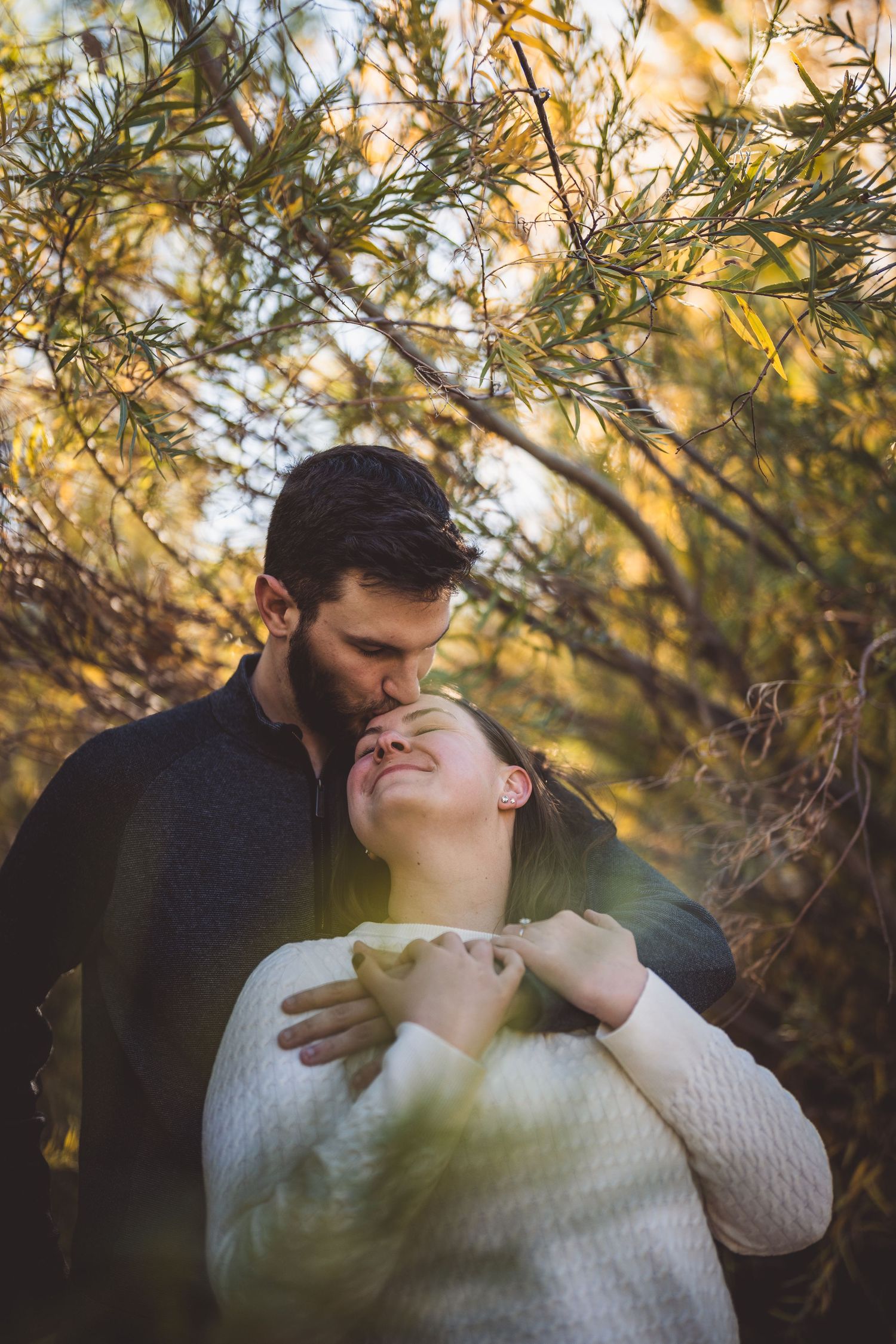 A couple shares a tender moment outdoors in autumn, man kissing womans forehead while embracing among golden foliage.