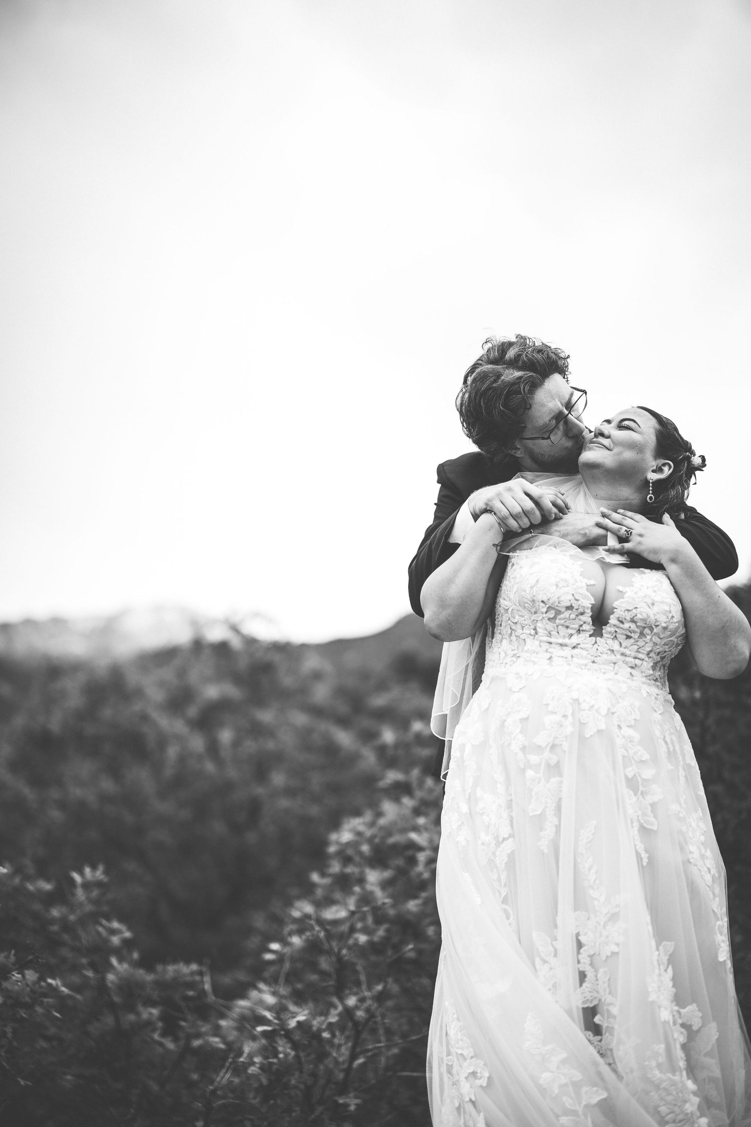 Black and white photo of a couple sharing a tender moment in their wedding attire against natural scenery.