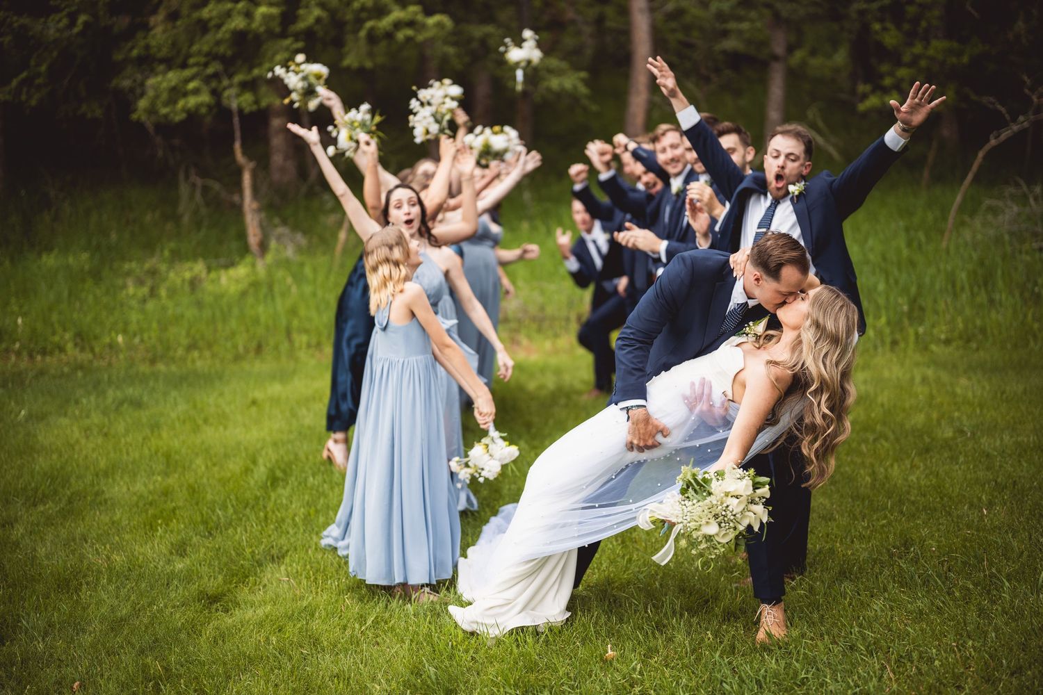 Wedding party posing joyfully on grassy lawn with bridesmaids in light blue dresses.