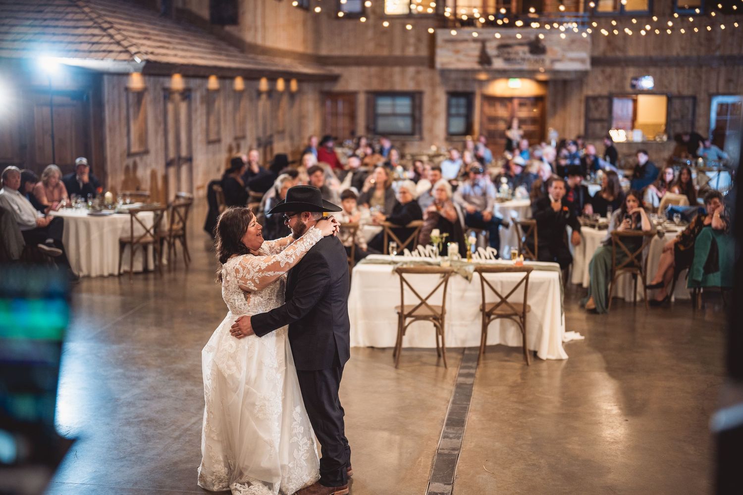 Wedding reception first dance in rustic barn venue with twinkling lights and guest tables.