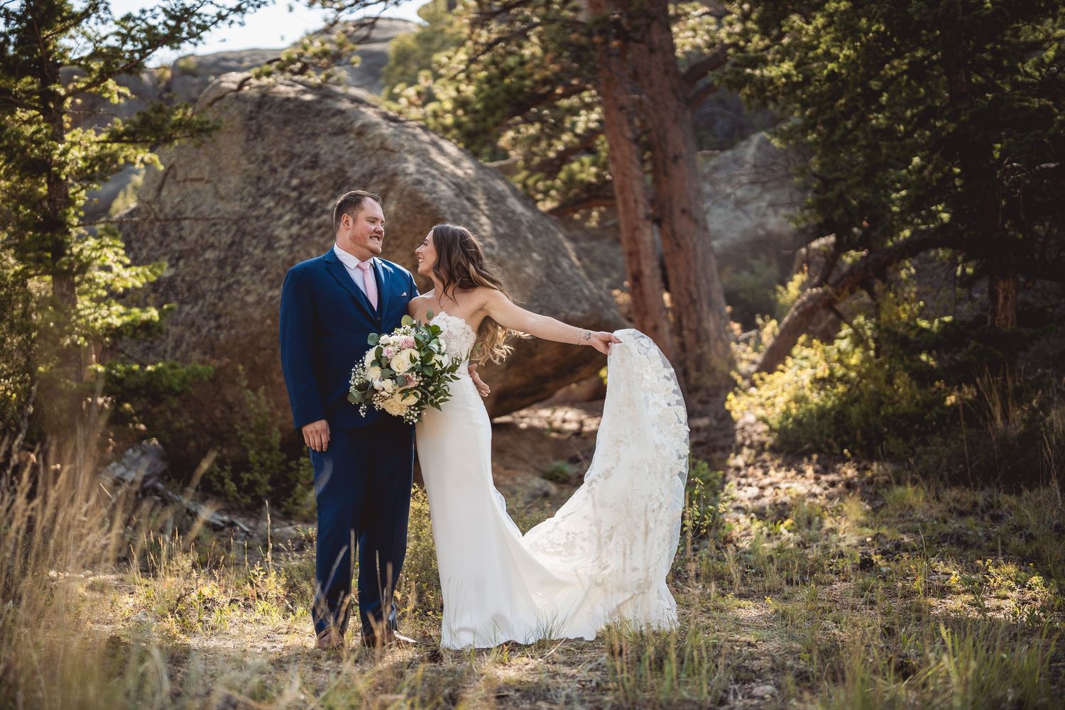 A newly married couple shares a romantic moment among tall pine trees in a rustic outdoor setting.