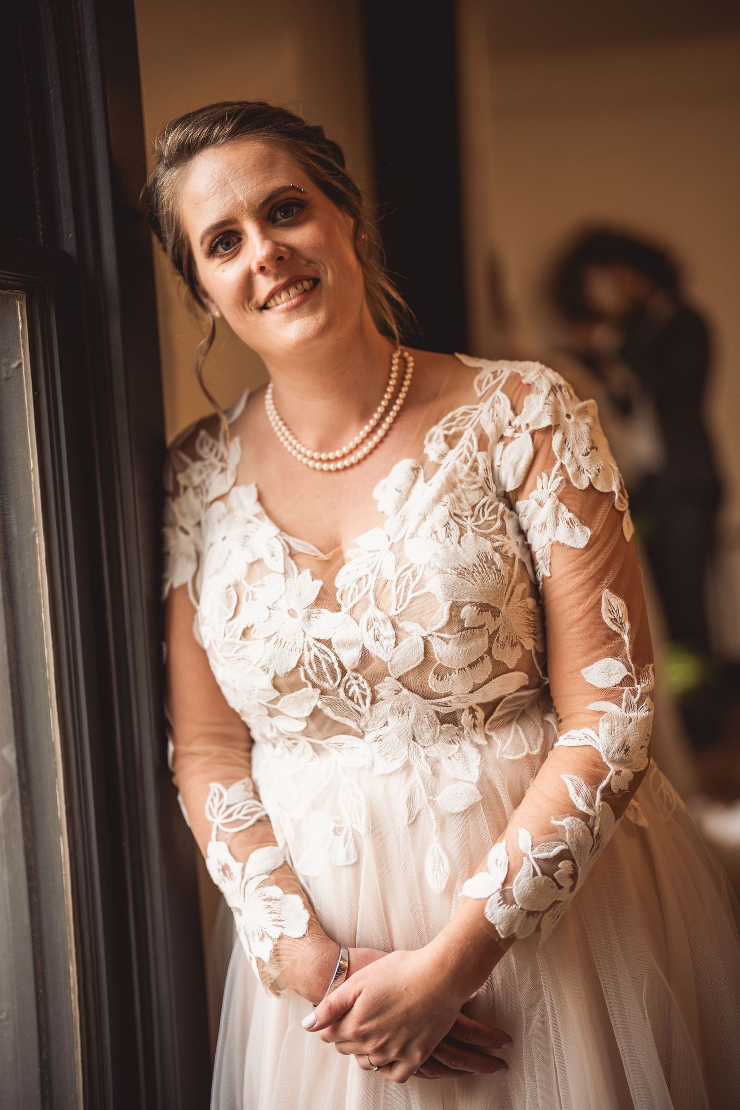 Bride in white lace gown with sheer sleeves smiles in natural window light wearing pearl necklace.