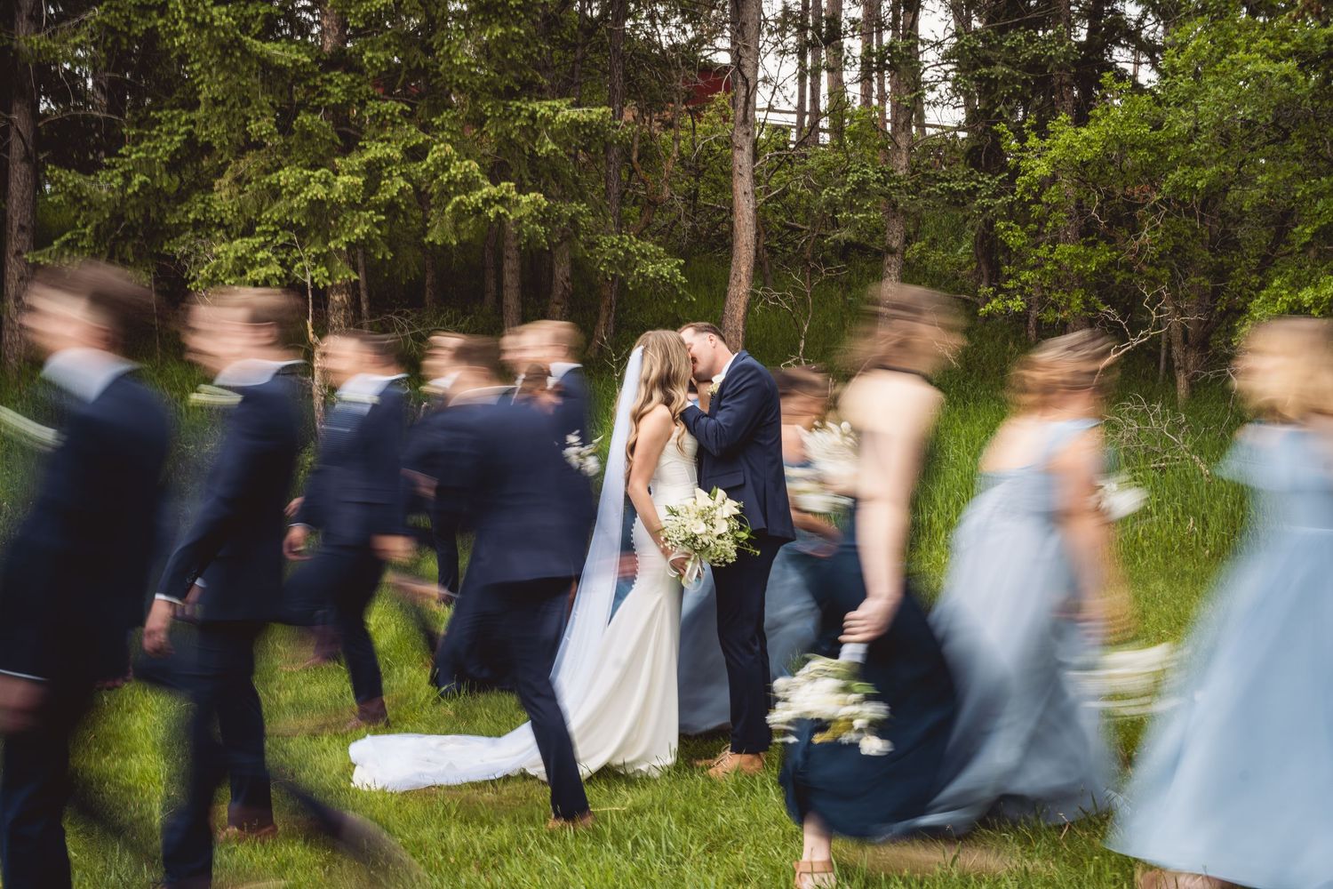 Bride and groom kiss while wedding party walks past in motion blur, outdoors near lush green forest.