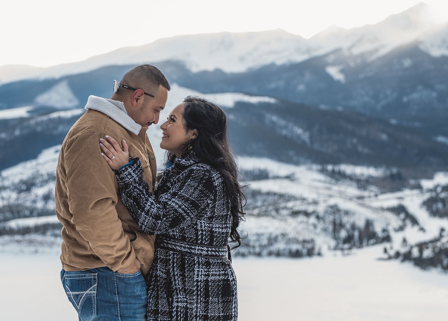 Couple embracing and gazing at each other with snowy mountain landscape backdrop in winter.